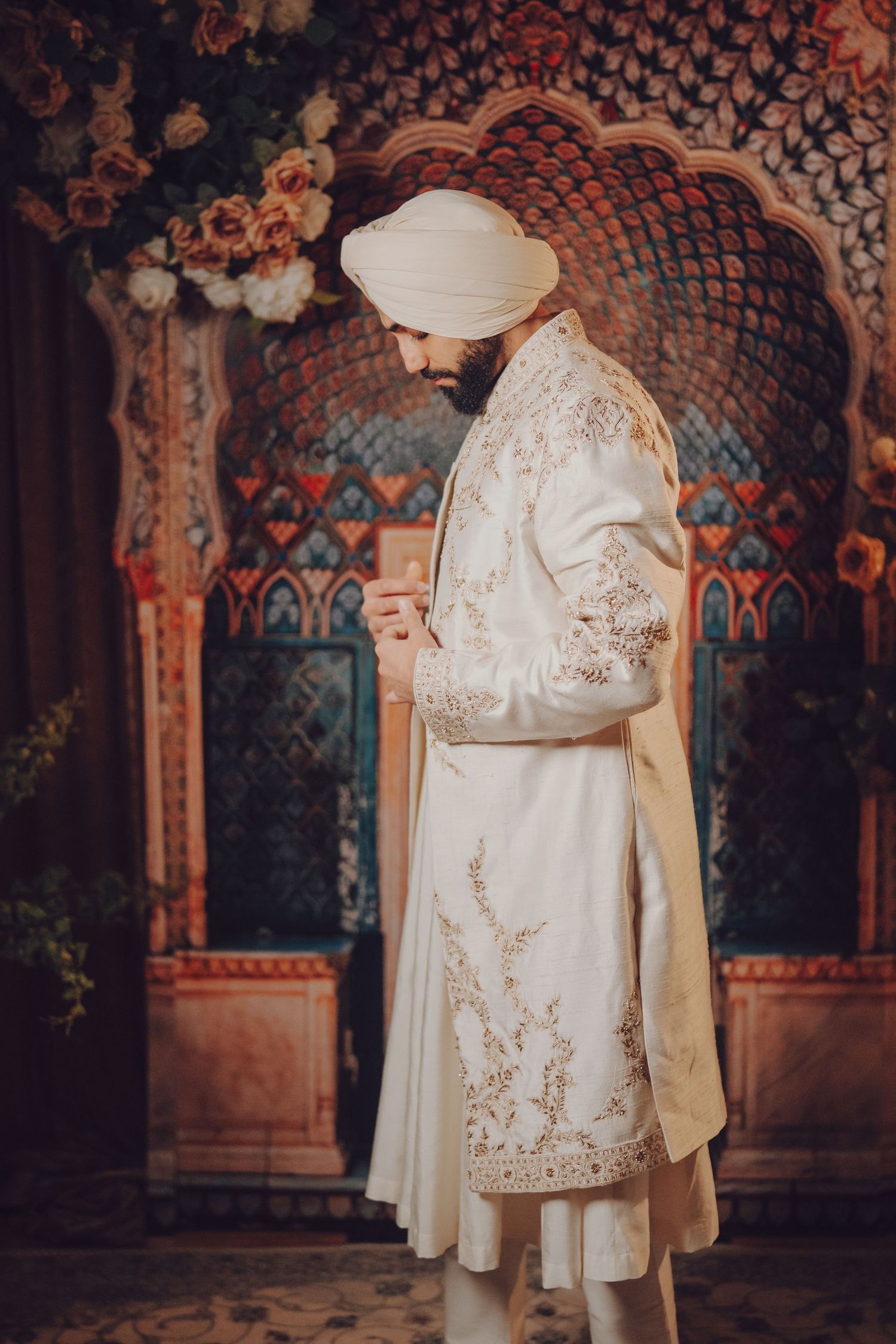 Traditional Sikh groom in white embroidered sherwani and turban stands in front of ornate archway.