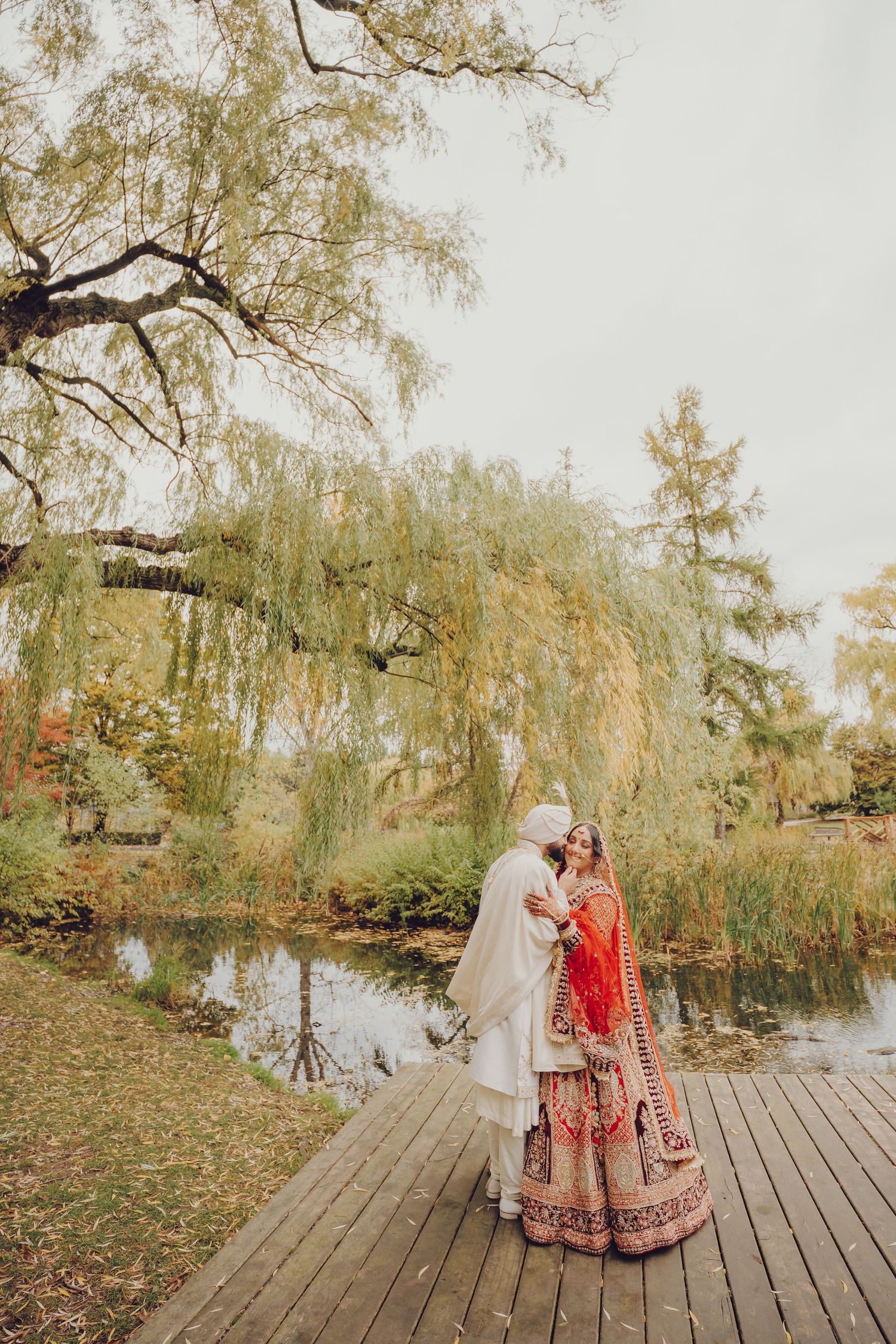 Traditional Indian wedding couple standing on wooden deck surrounded by willow trees.