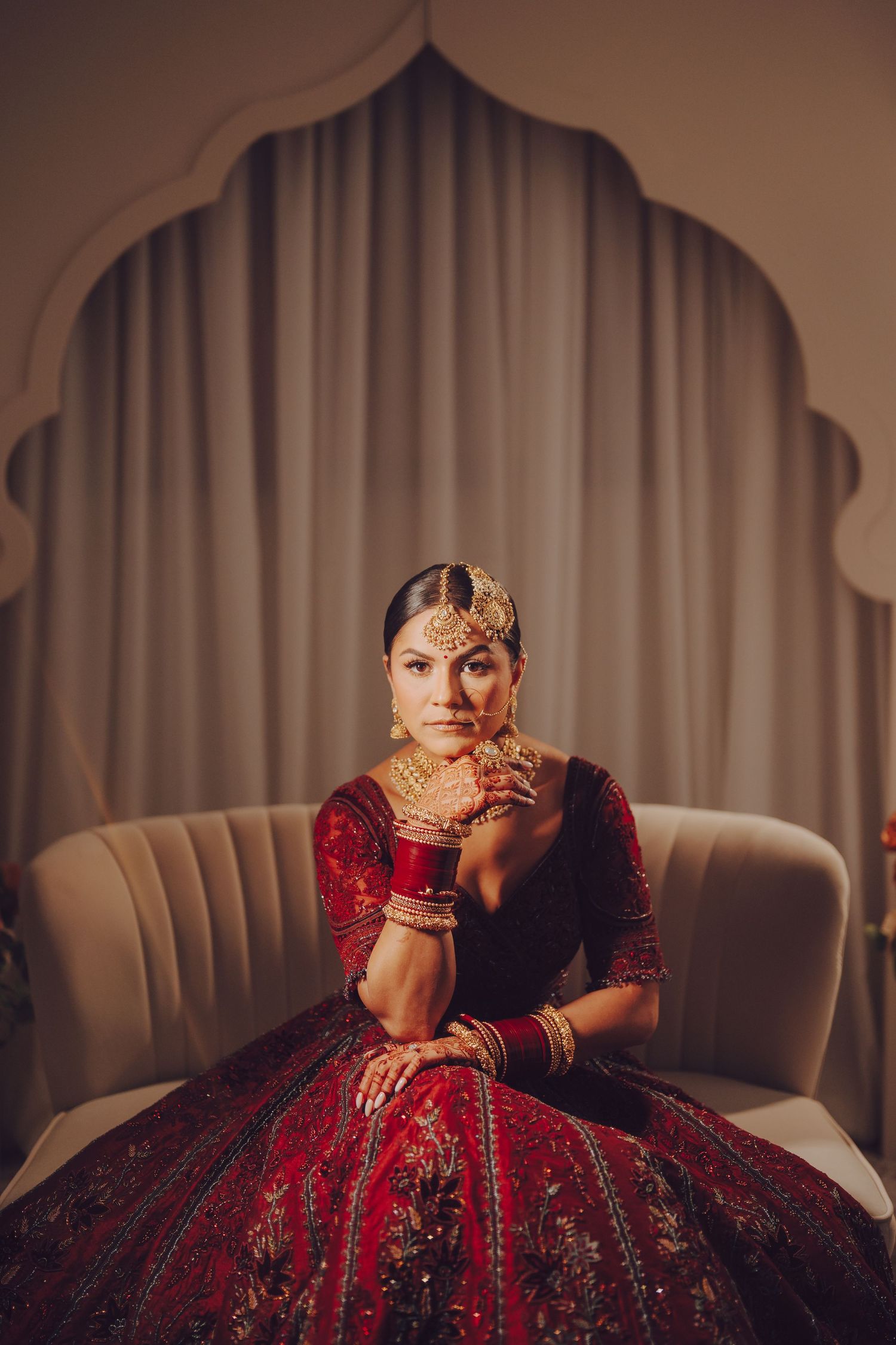 Elegant bridal portrait series showing a red and gold lehenga on a beige couch with dramatic lighting.