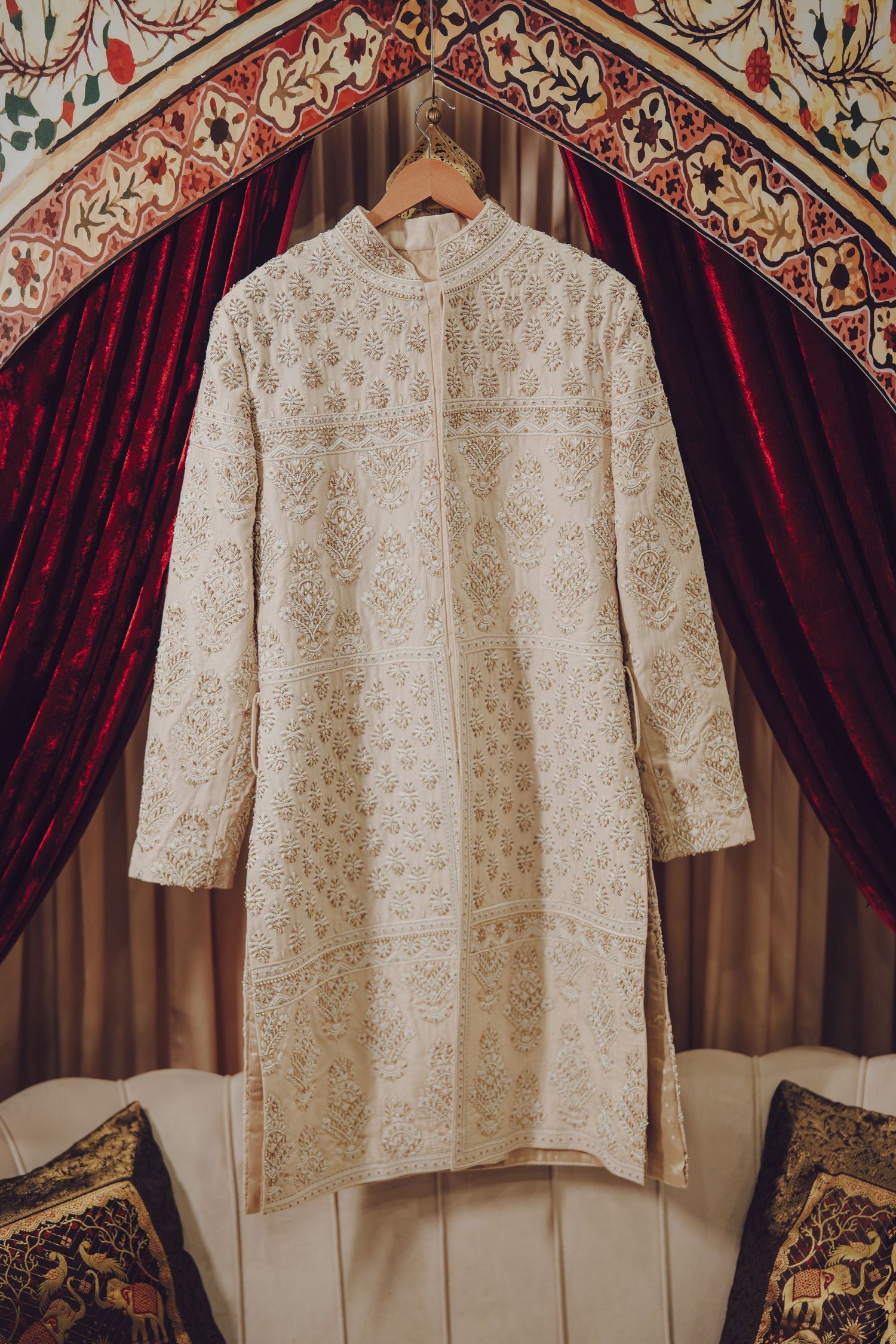Traditional white embroidered sherwani displayed against an ornate backdrop with red velvet curtains.