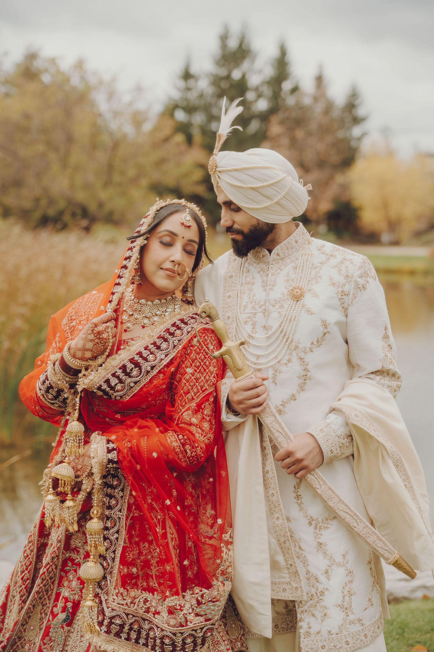 A couple in traditional South Asian wedding attire poses outdoors in autumn, with the bride in red and groom in white.