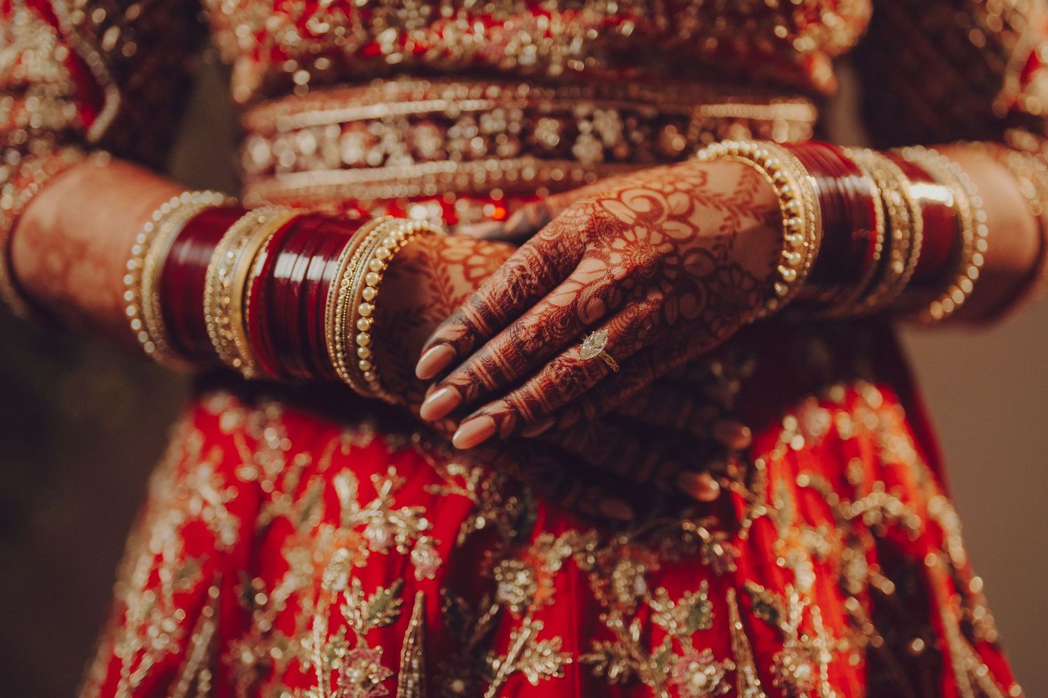 Traditional Indian bridal hands adorned with henna designs and red bangles against a red and gold wedding outfit.