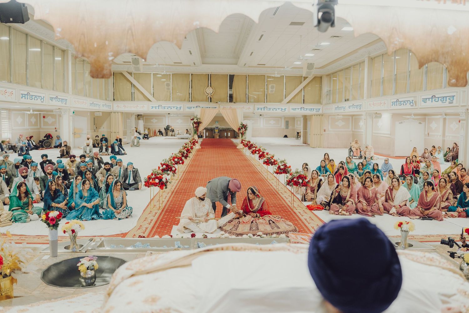 Panoramic view of a Sikh wedding ceremony with guests seated on the floor in a decorated hall.