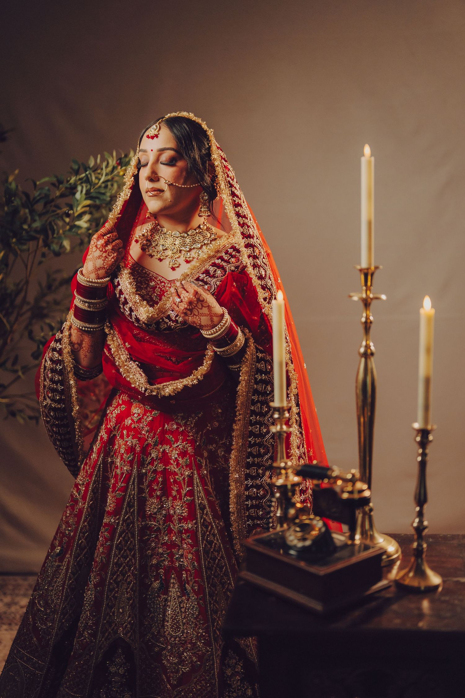 Traditional Indian bride in ornate red lehenga poses elegantly with lit candles in moody lighting.