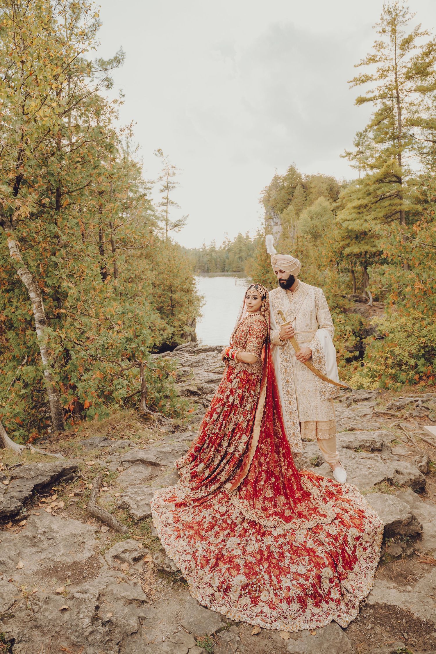 A couple in traditional Indian wedding attire stands on rocky terrain with a lake and trees in the background.