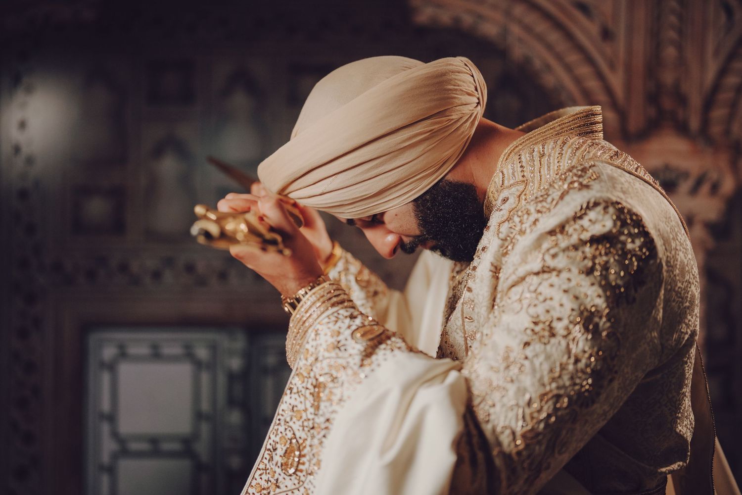 Groom in traditional cream and gold Sikh wedding attire adjusting turban.