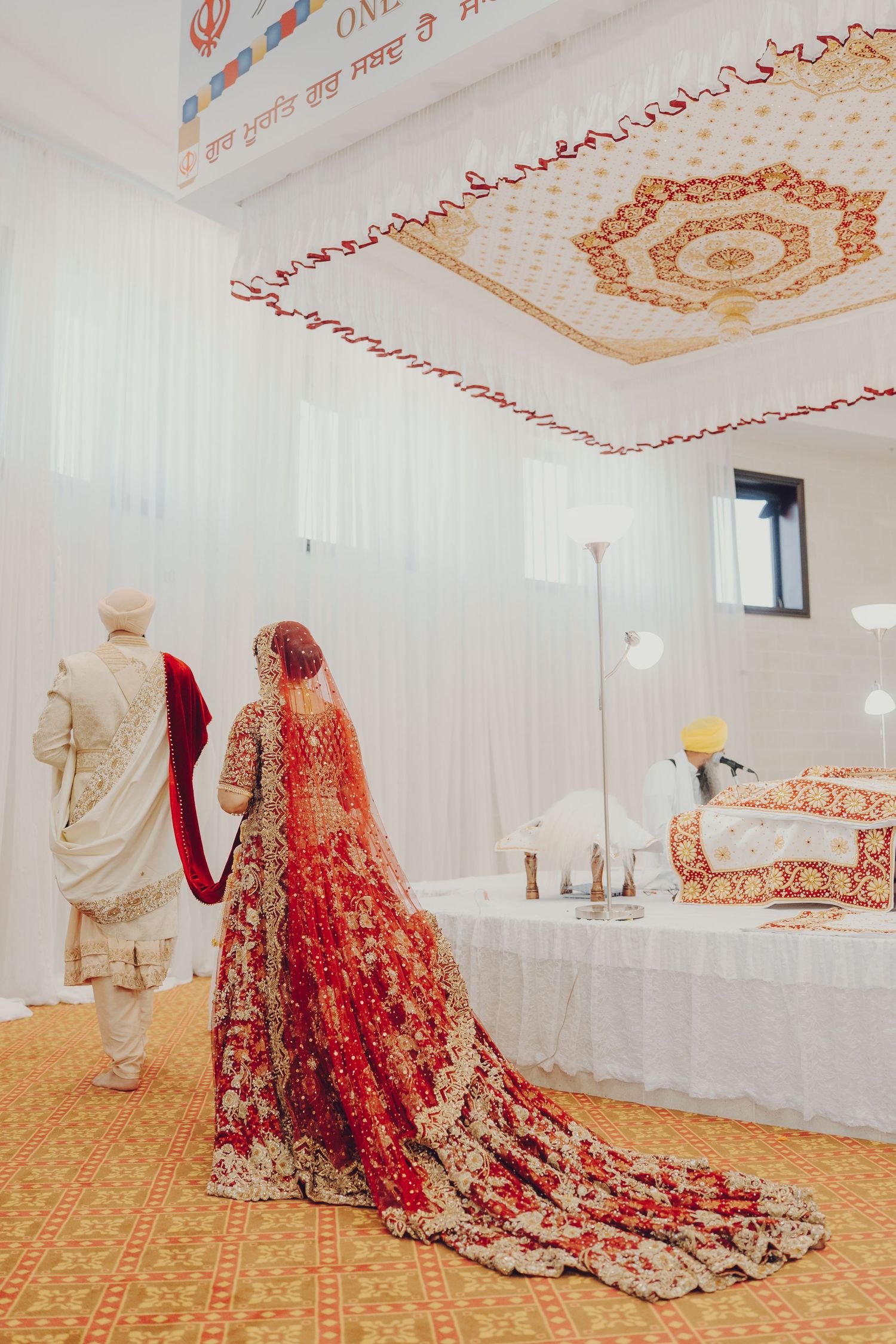 Traditional red and gold embroidered bridal lehenga displayed in an elegant white ceremonial room with decorative ceiling.