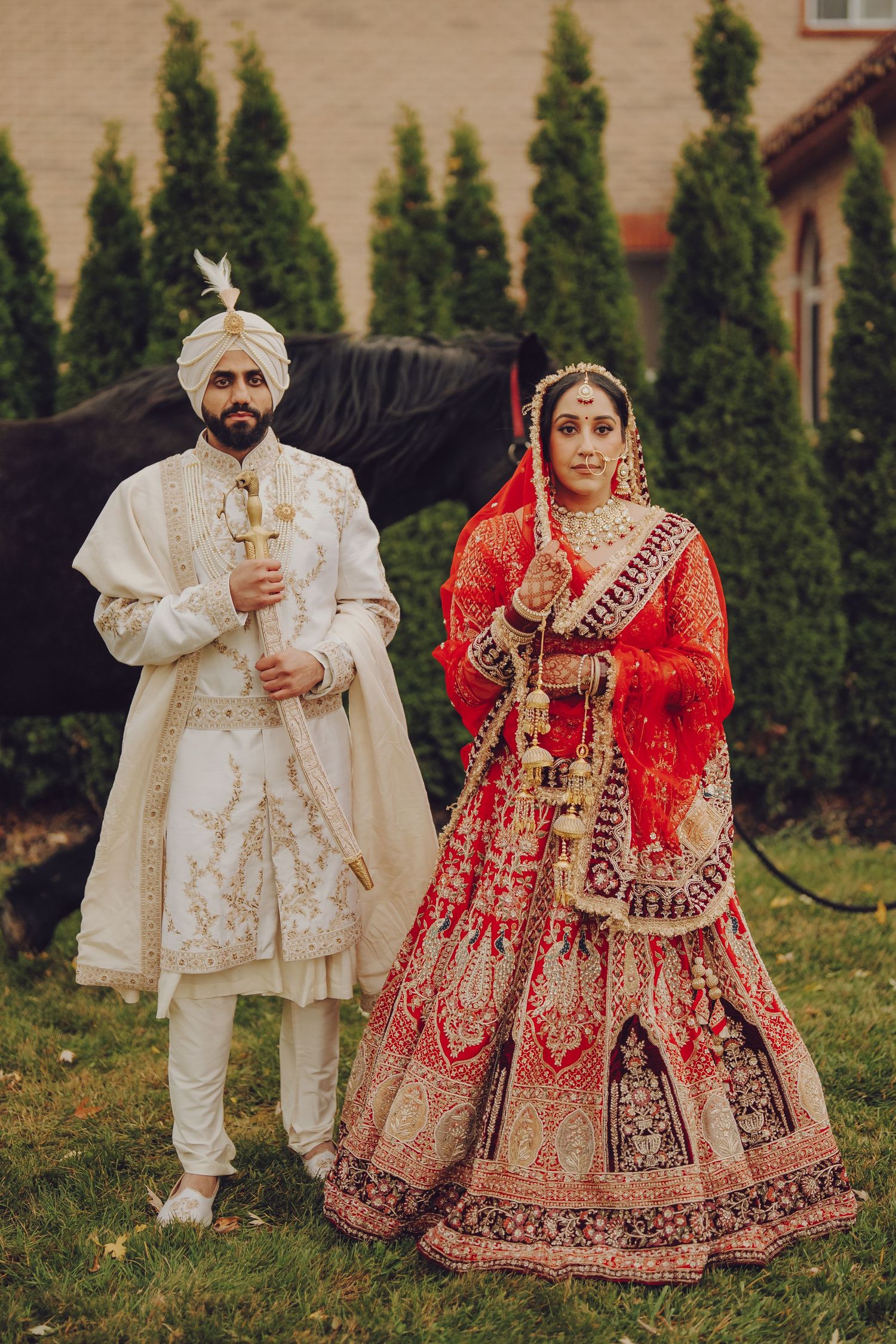 Wedding couple in traditional Sikh attire posing in garden with evergreen trees.