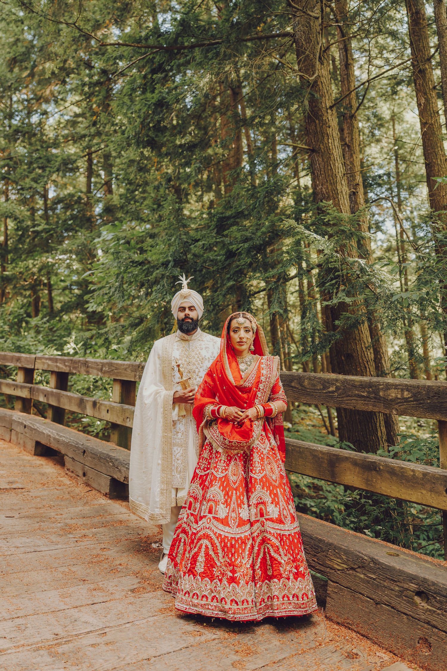 Wedding couple in traditional Indian attire posing on wooden boardwalk surrounded by tall evergreen trees.