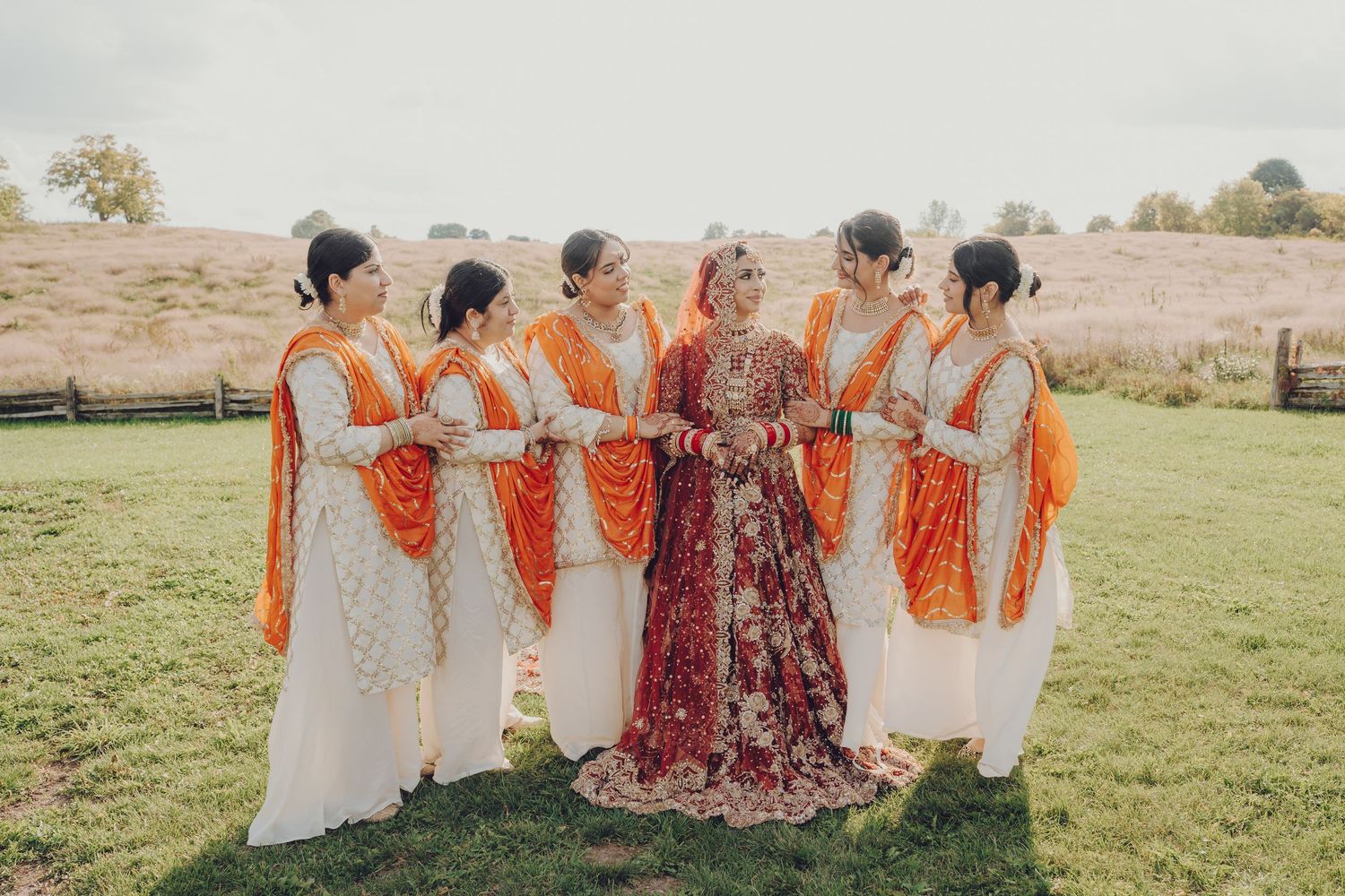 Wedding party in orange and burgundy dresses poses together in a sunny field.