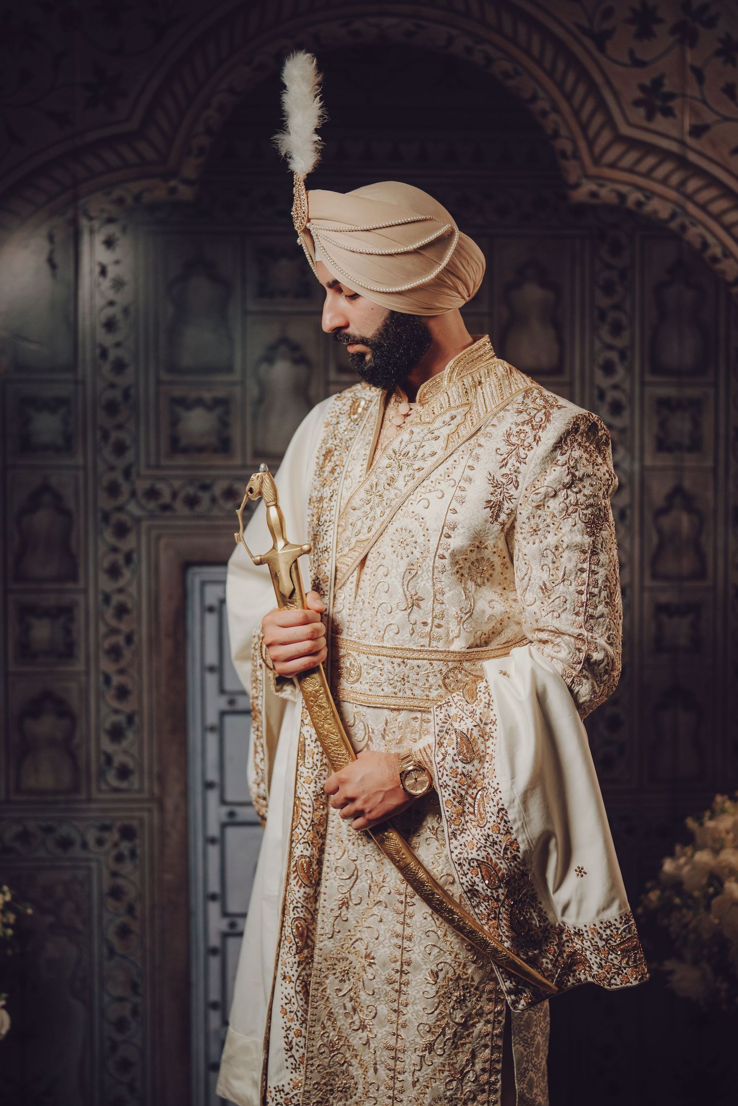 Person in cream and gold embroidered attire holding ceremonial sword against decorative backdrop.