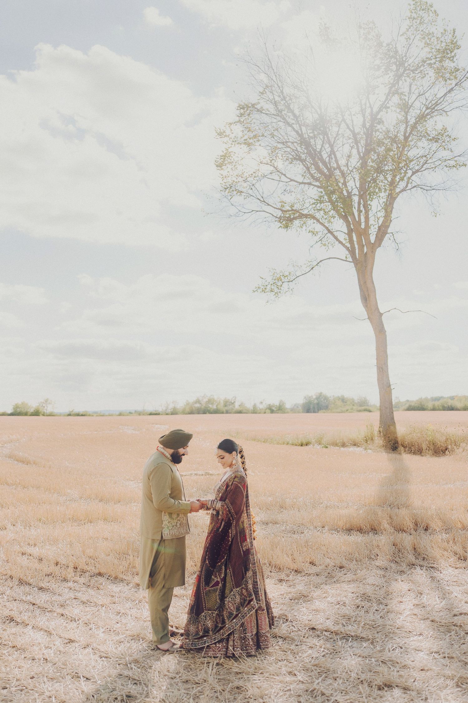 Couple in traditional South Asian attire poses together in a sunlit field with a lone tree in the background.