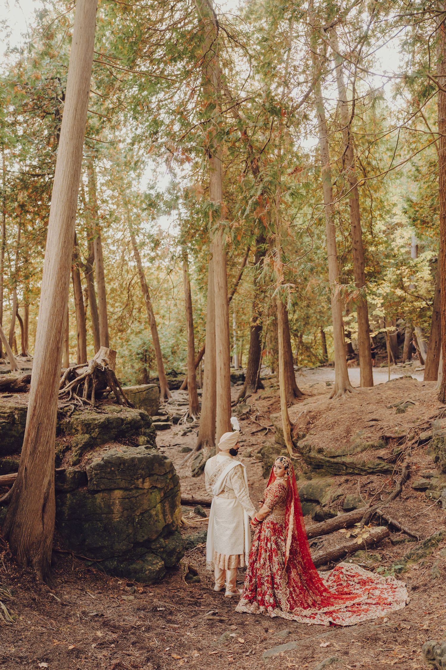 A couple in traditional wedding attire walks through a scenic wooded forest path.
