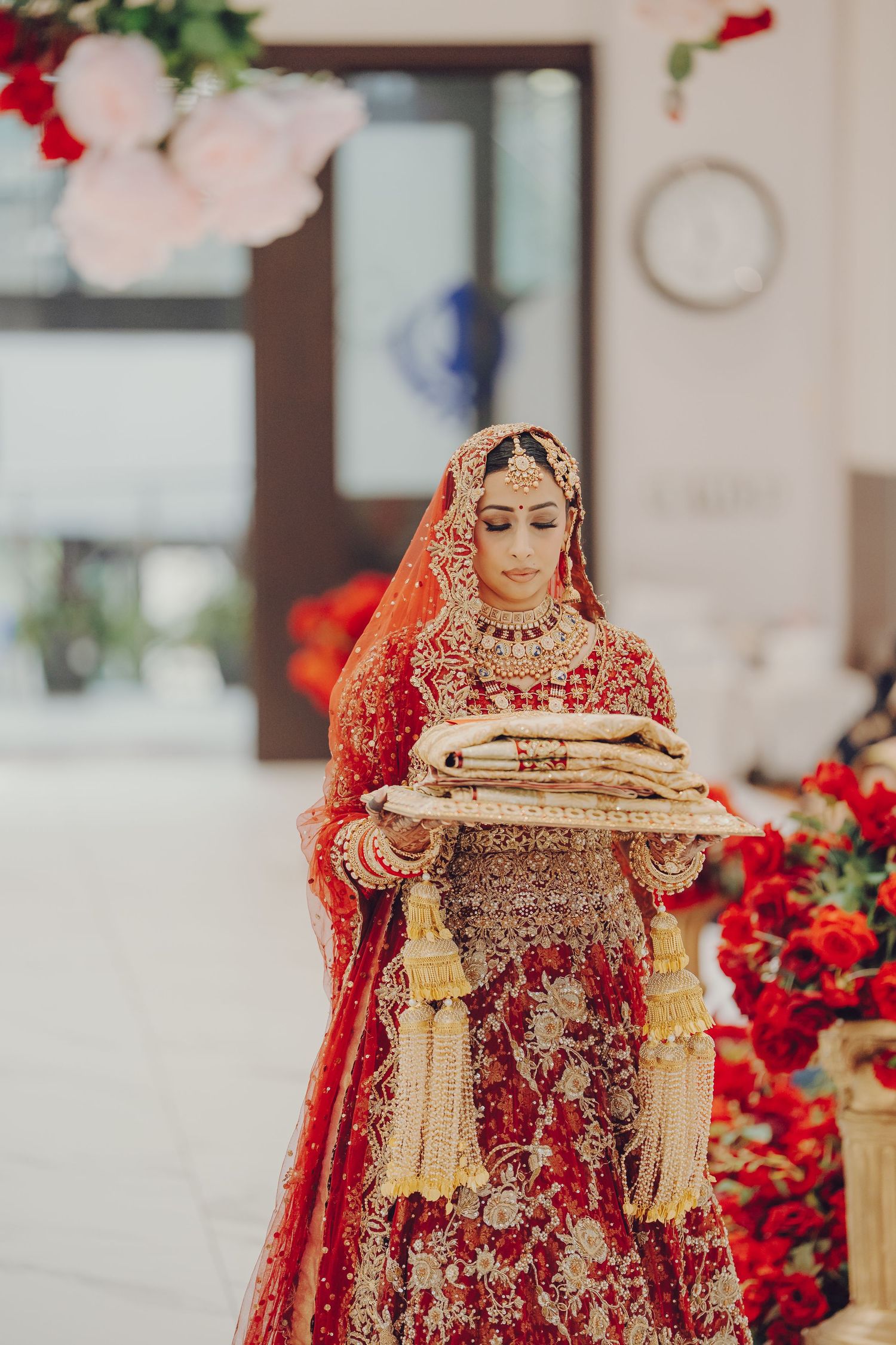 Traditional Indian bride in ornate red lehenga with gold embroidery and jewelry stands amid floral decor.
