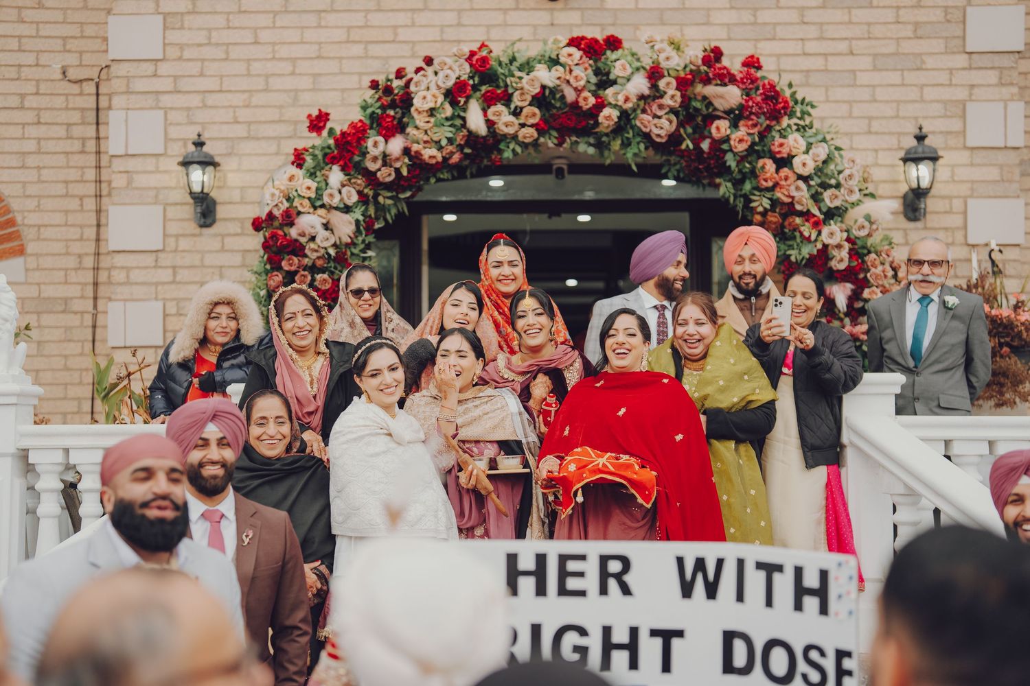 Wedding party gathered under a floral arch with red roses against a brick wall backdrop.