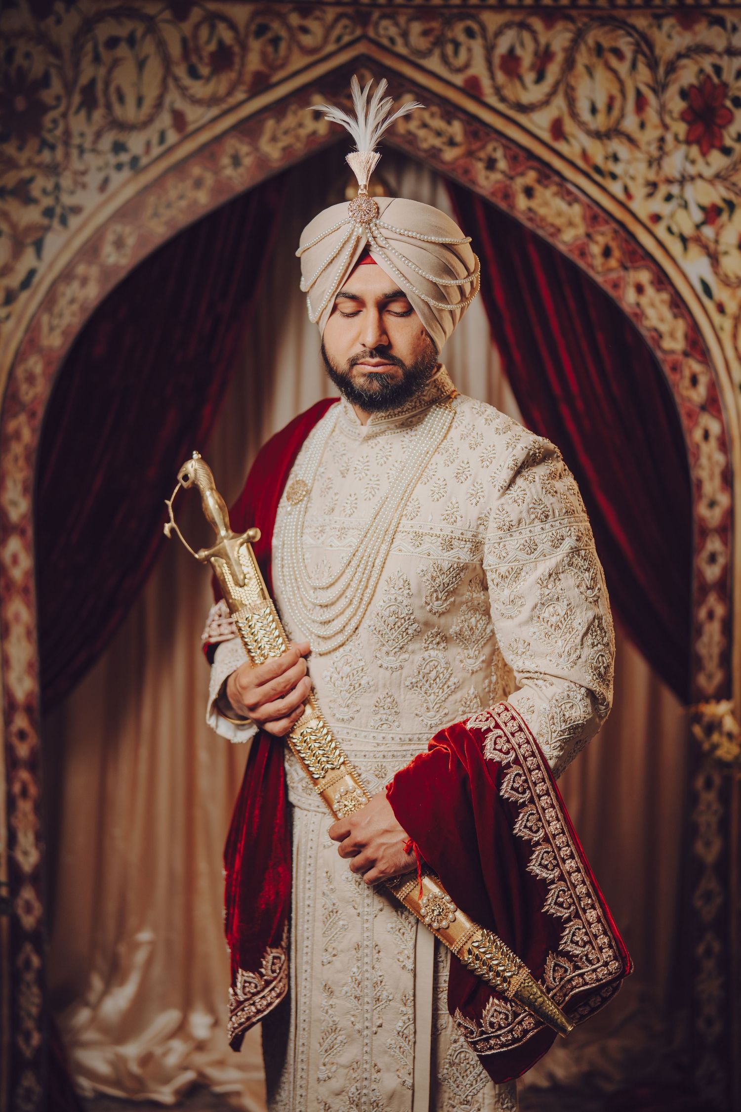 Groom in cream sherwani and turban holds ceremonial sword against ornate backdrop.