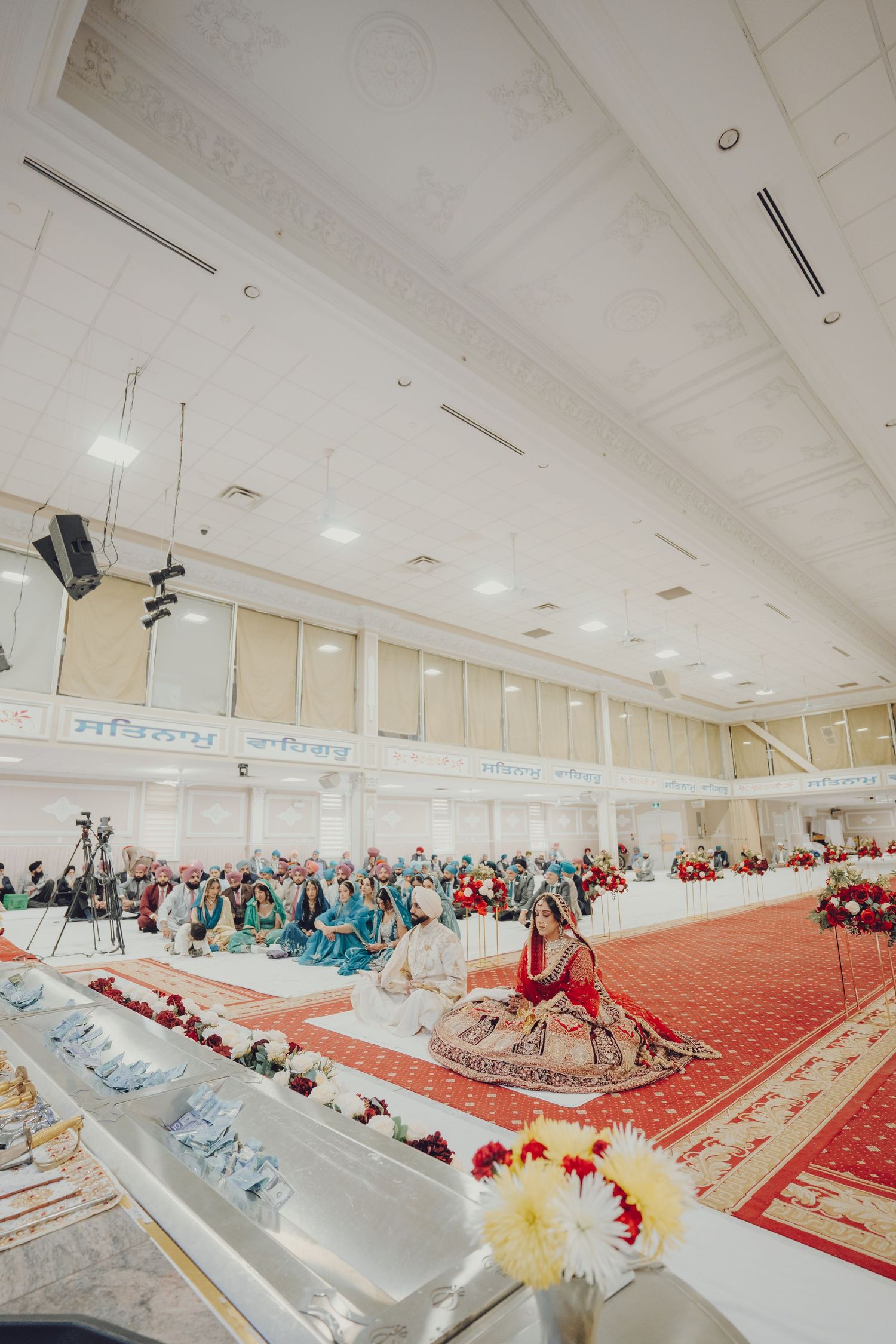 Wide view of a Sikh wedding ceremony in a decorated hall with guests seated on the floor.
