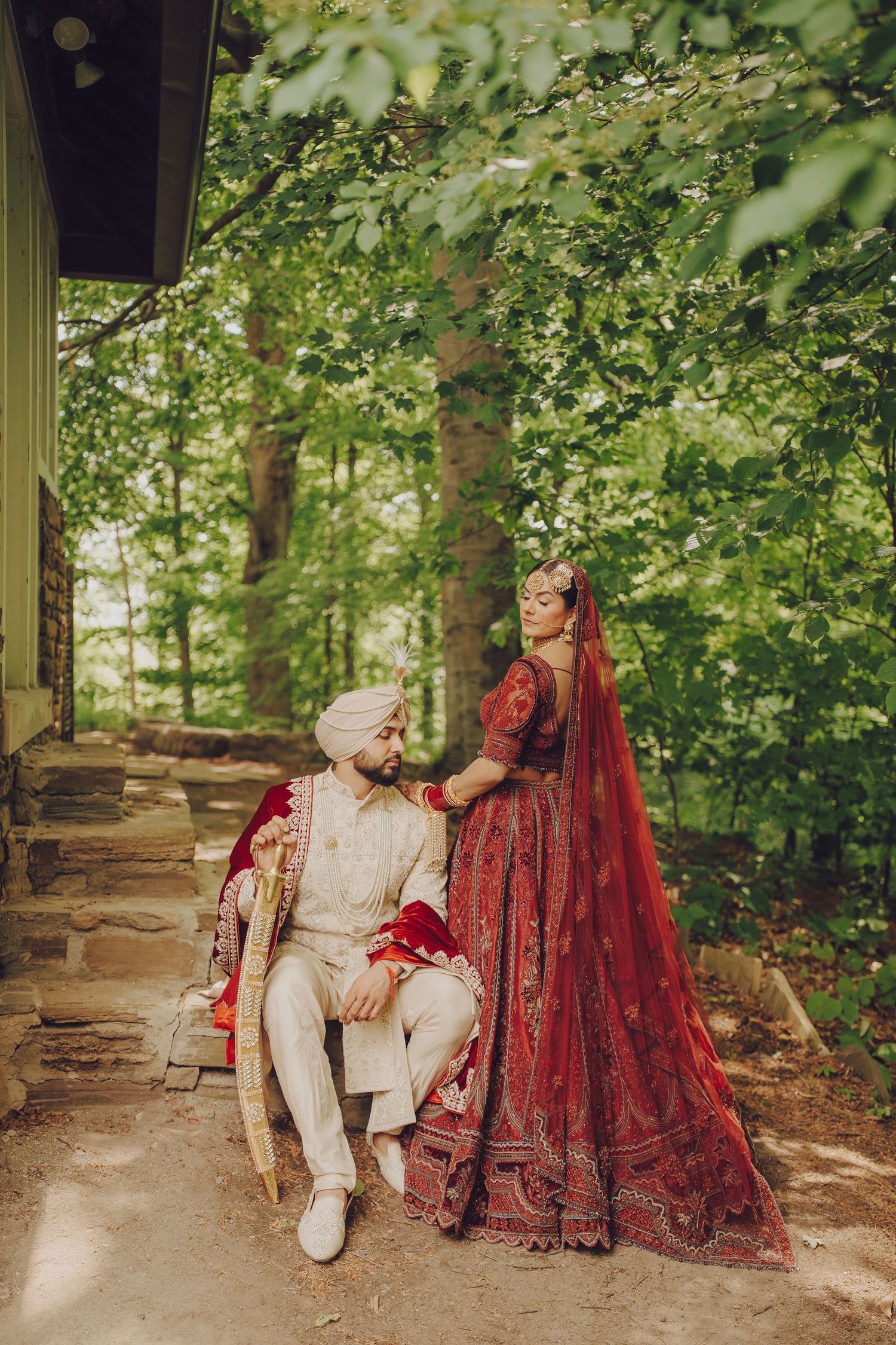 Couple in traditional Indian wedding dress share tender moments on stone benches surrounded by lush greenery.