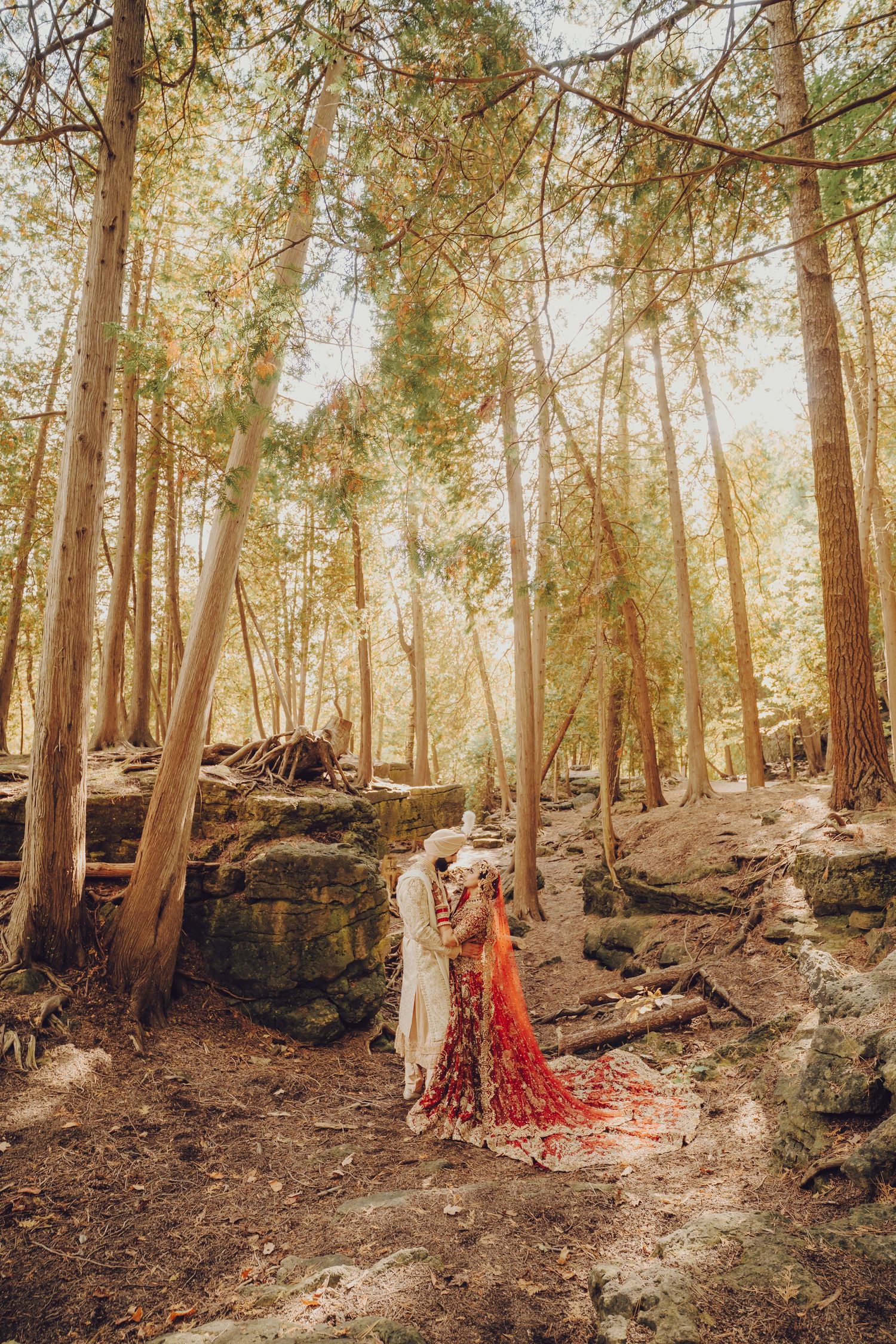 A wedding couple in traditional attire poses among tall trees in a forest setting.