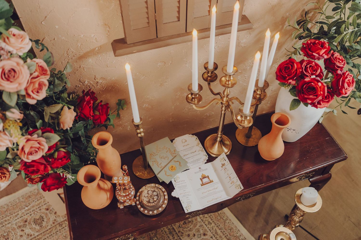 Elegant table setting with white candles, terracotta vases, and pink and red rose floral arrangements on wooden surface.