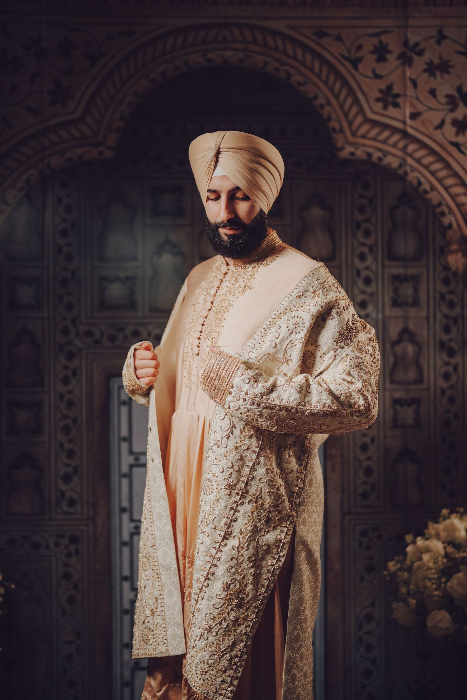 Traditional Sikh groom wearing cream sherwani poses against intricate architectural backdrop.