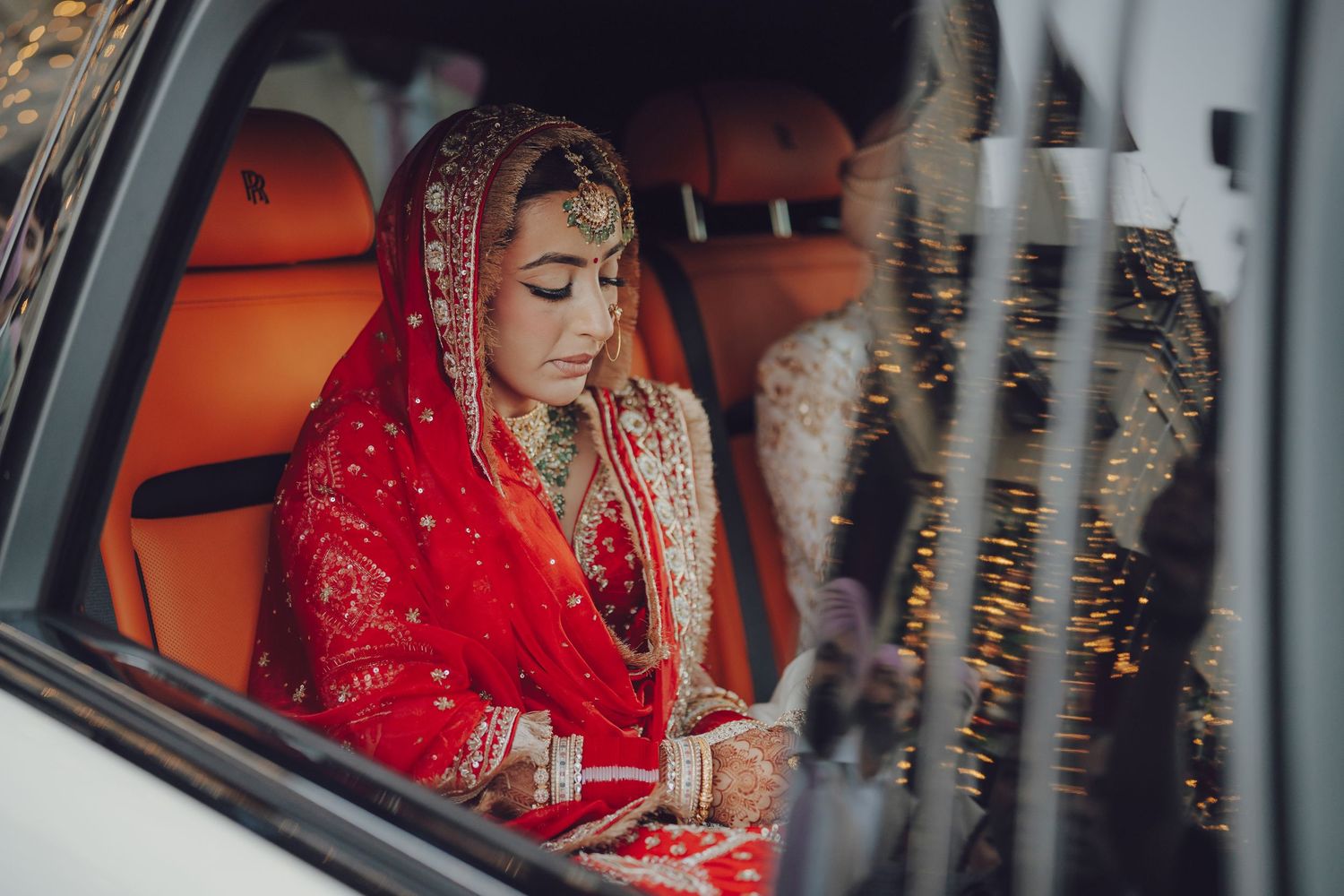 Bride in red traditional Indian wedding attire seated in car with intricate jewelry and details.