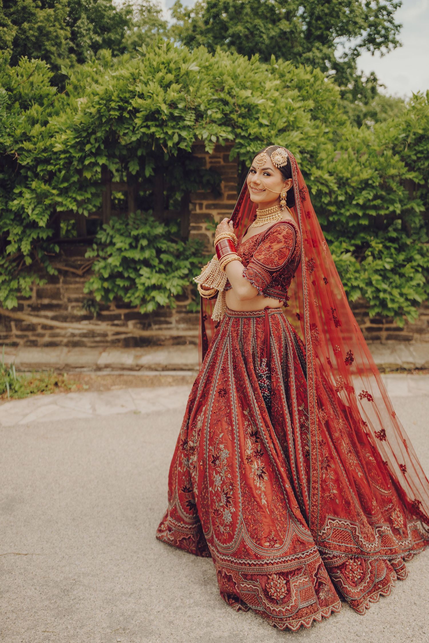 Traditional South Asian bridal attire in rich red and gold embroidered lehenga with flowing veil captured in outdoor setting.