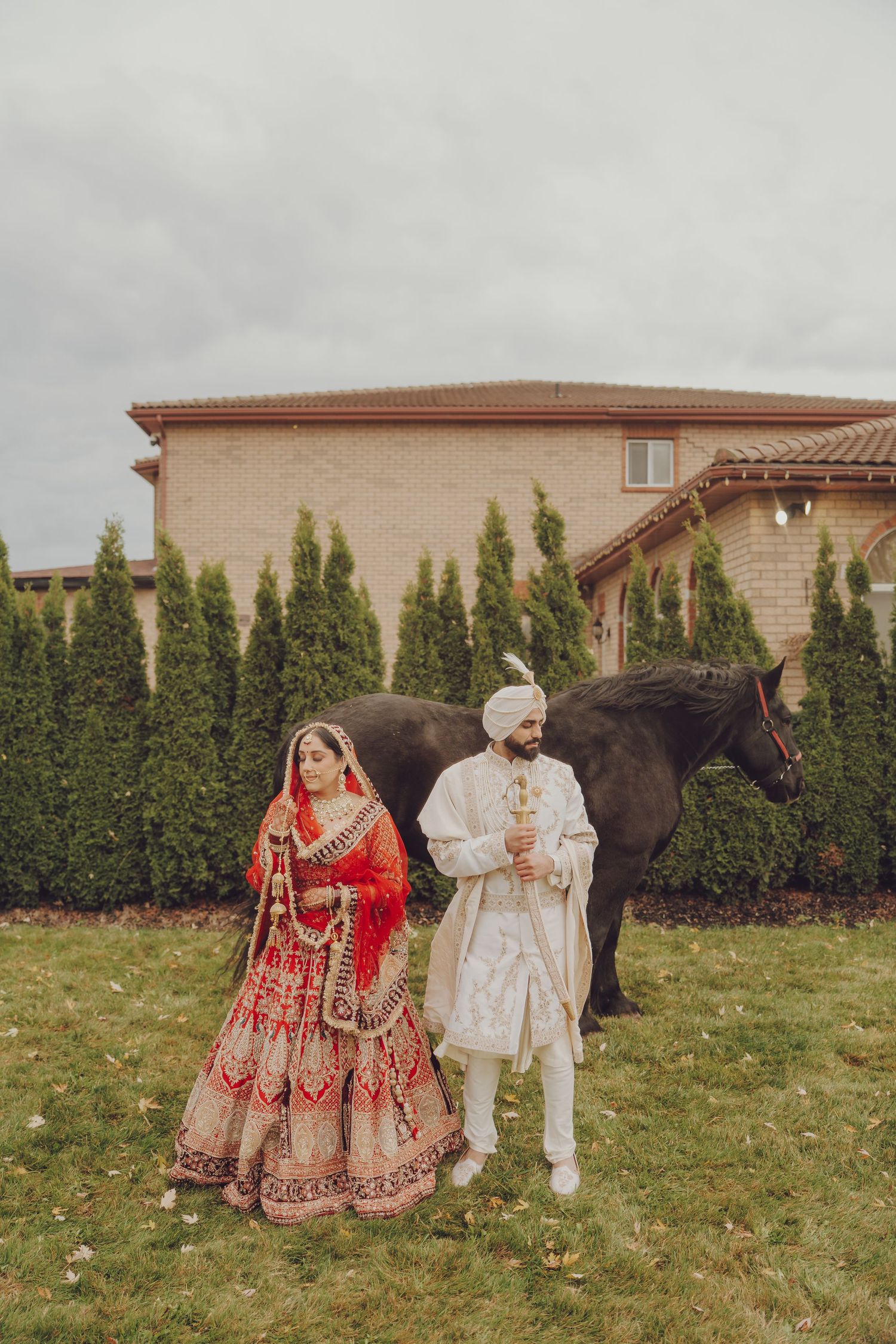 A couple in traditional South Asian wedding attire poses together outdoors against a backdrop of evergreen trees.