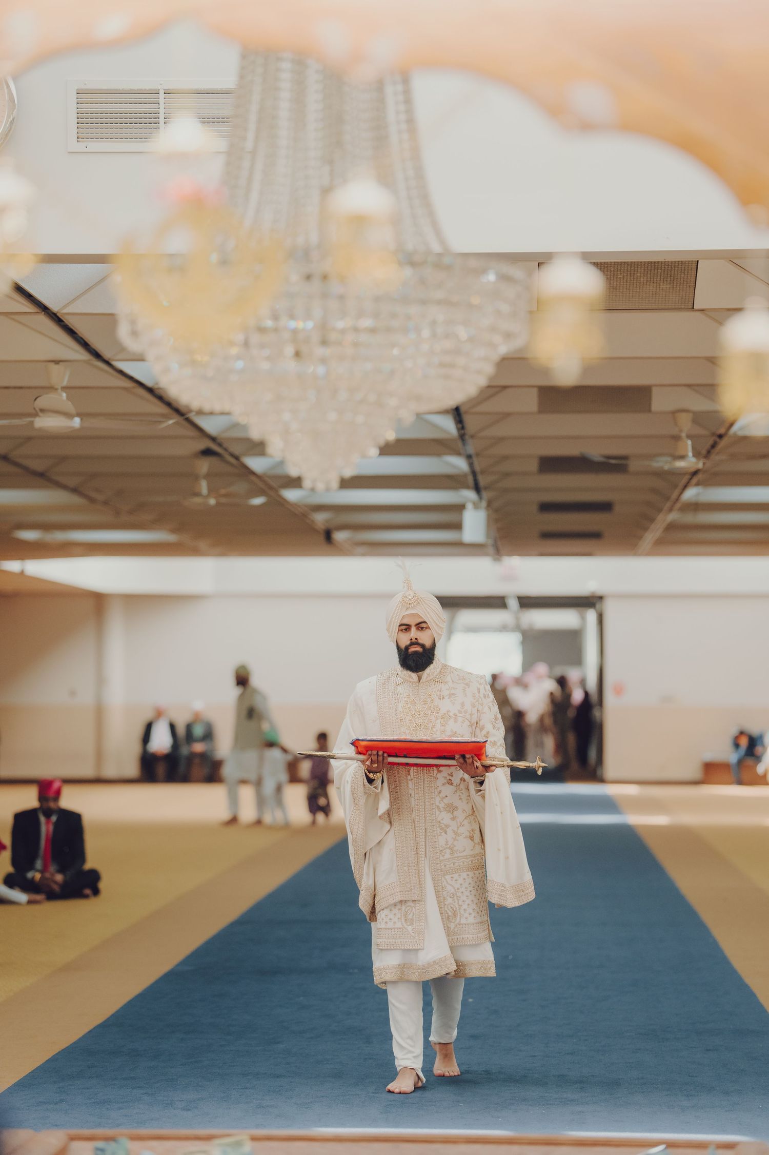 Traditional Sikh wedding ceremony taking place on a blue carpeted floor with ornate chandeliers overhead.