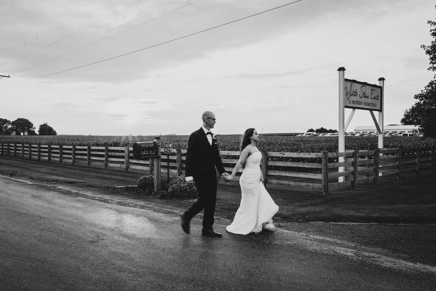 Black and white photo of a bride and groom holding hands while crossing a wet road near a wooden fence. White Stone Events Sugar Grove IL
