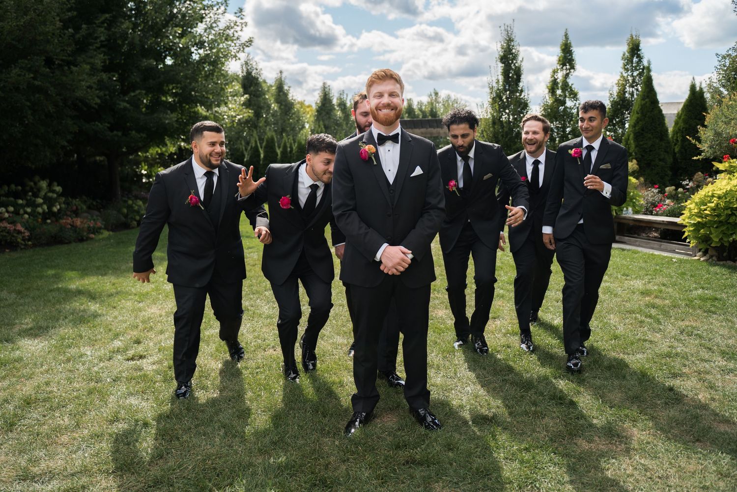 Groom in black tuxedo with groomsmen in matching suits and red boutonnieres walking outdoors.