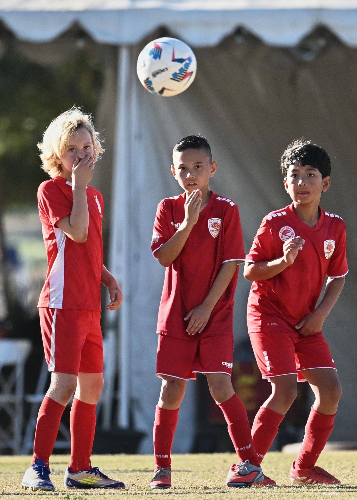 Three players in red uniforms watch a soccer ball float above their heads.