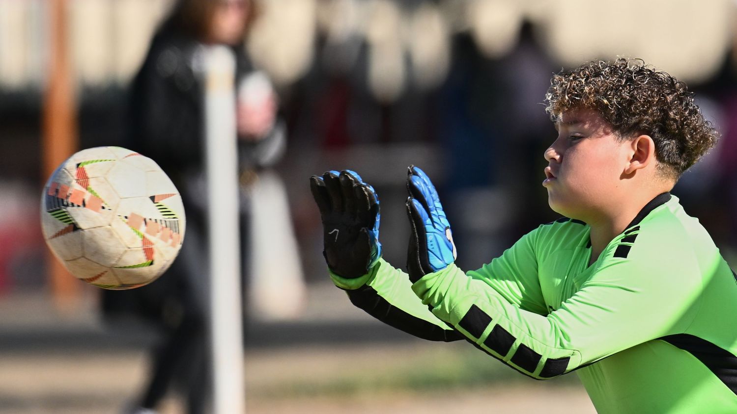 A youth soccer goalkeeper in a neon green jersey reaches to catch a soccer ball during a match.