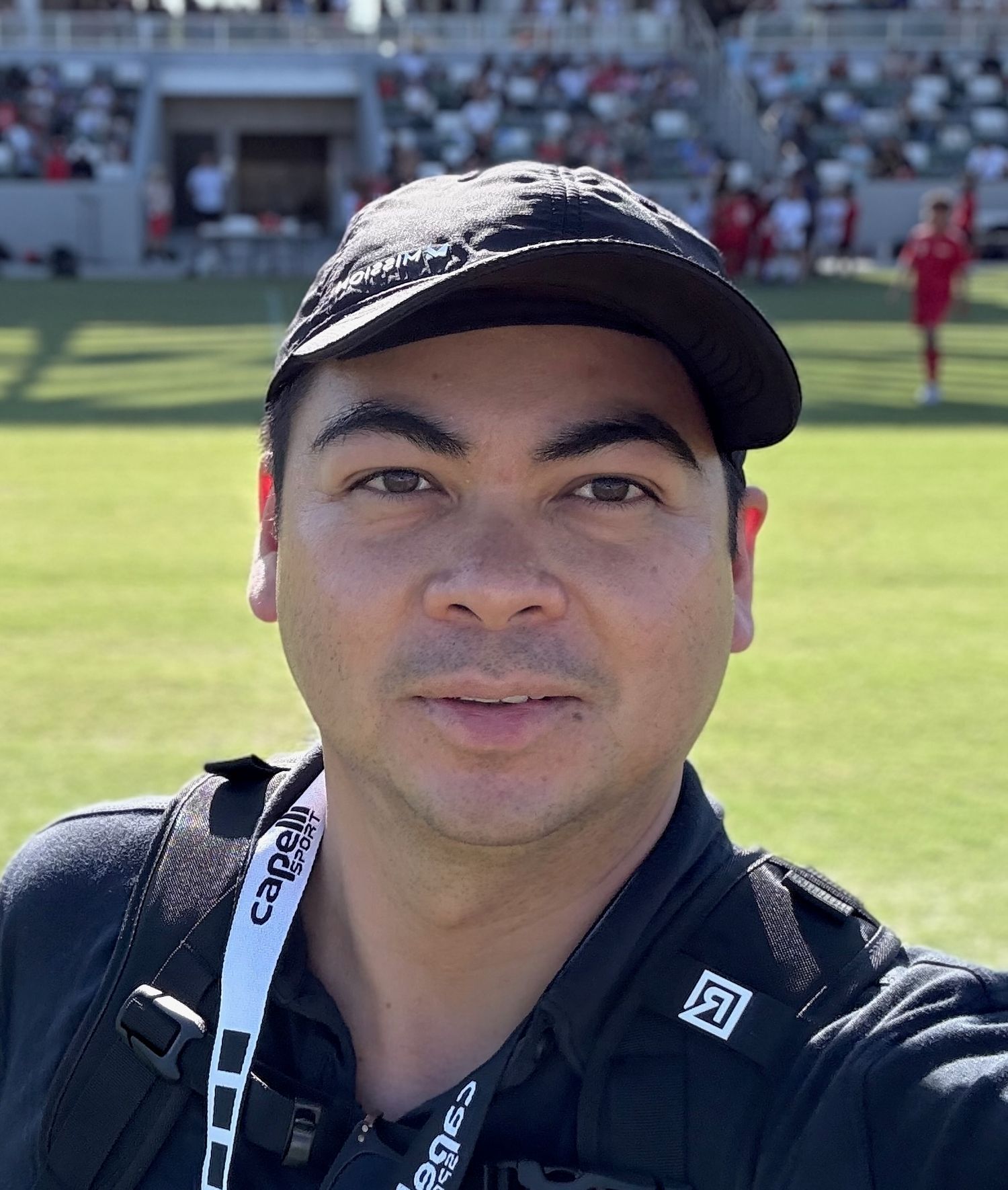 Person wearing a black baseball cap and lanyard smiling at a sporting event.