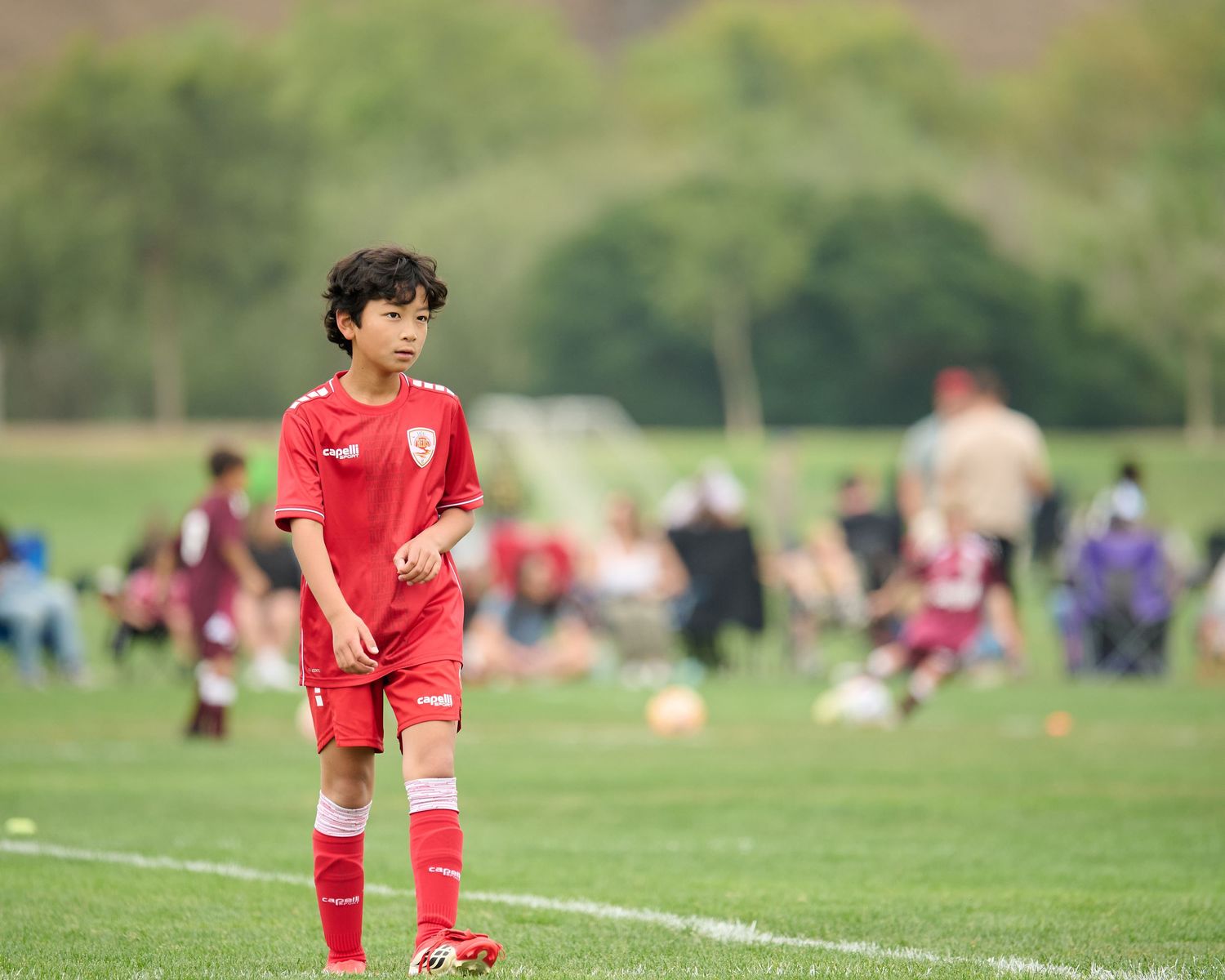 Young boy in red soccer uniform playing on a green field during a youth soccer match outdoors.