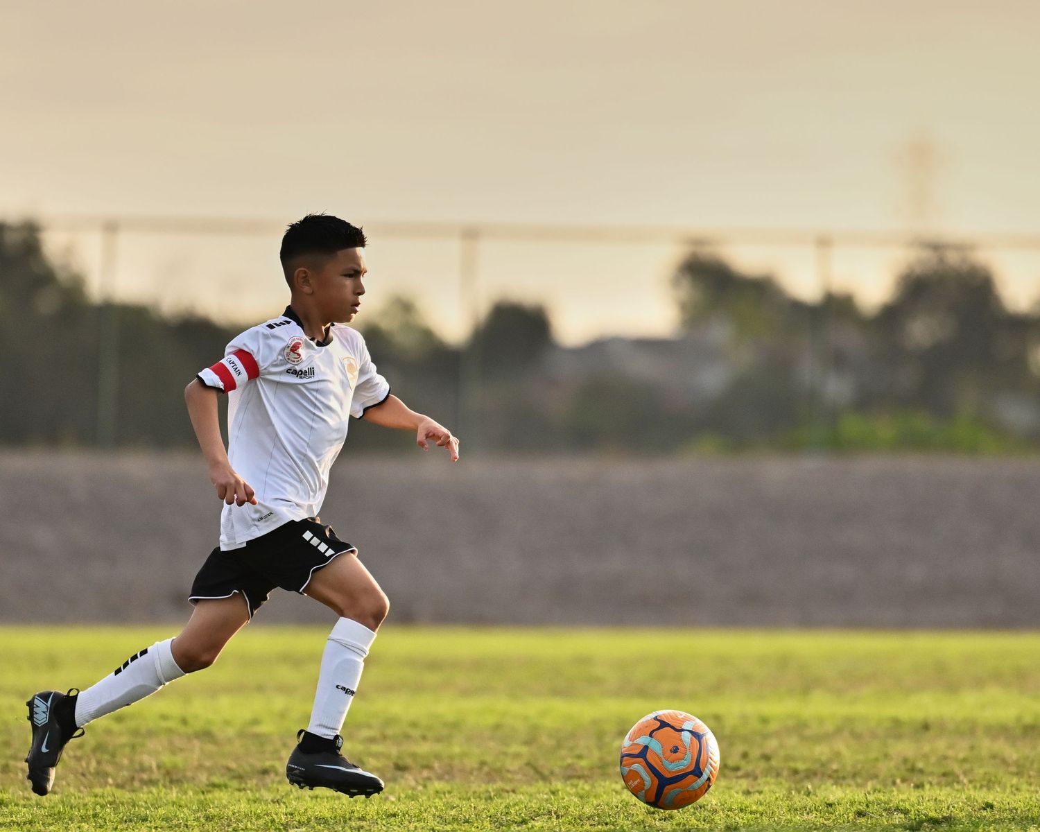 Soccer player in white uniform running with the ball during sunset on a grass field.