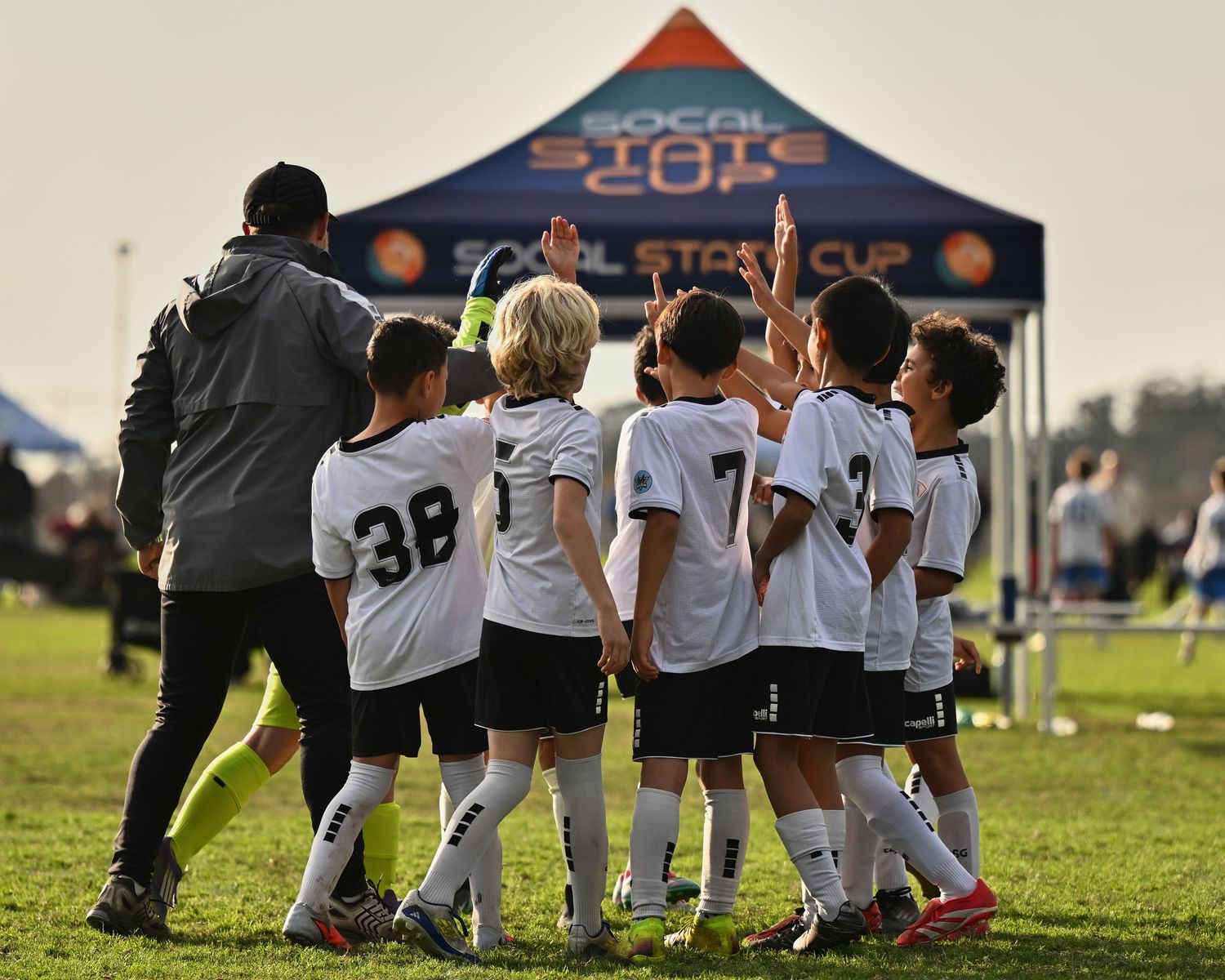 Youth soccer team huddles with their coach under a blue tent on a grass field.