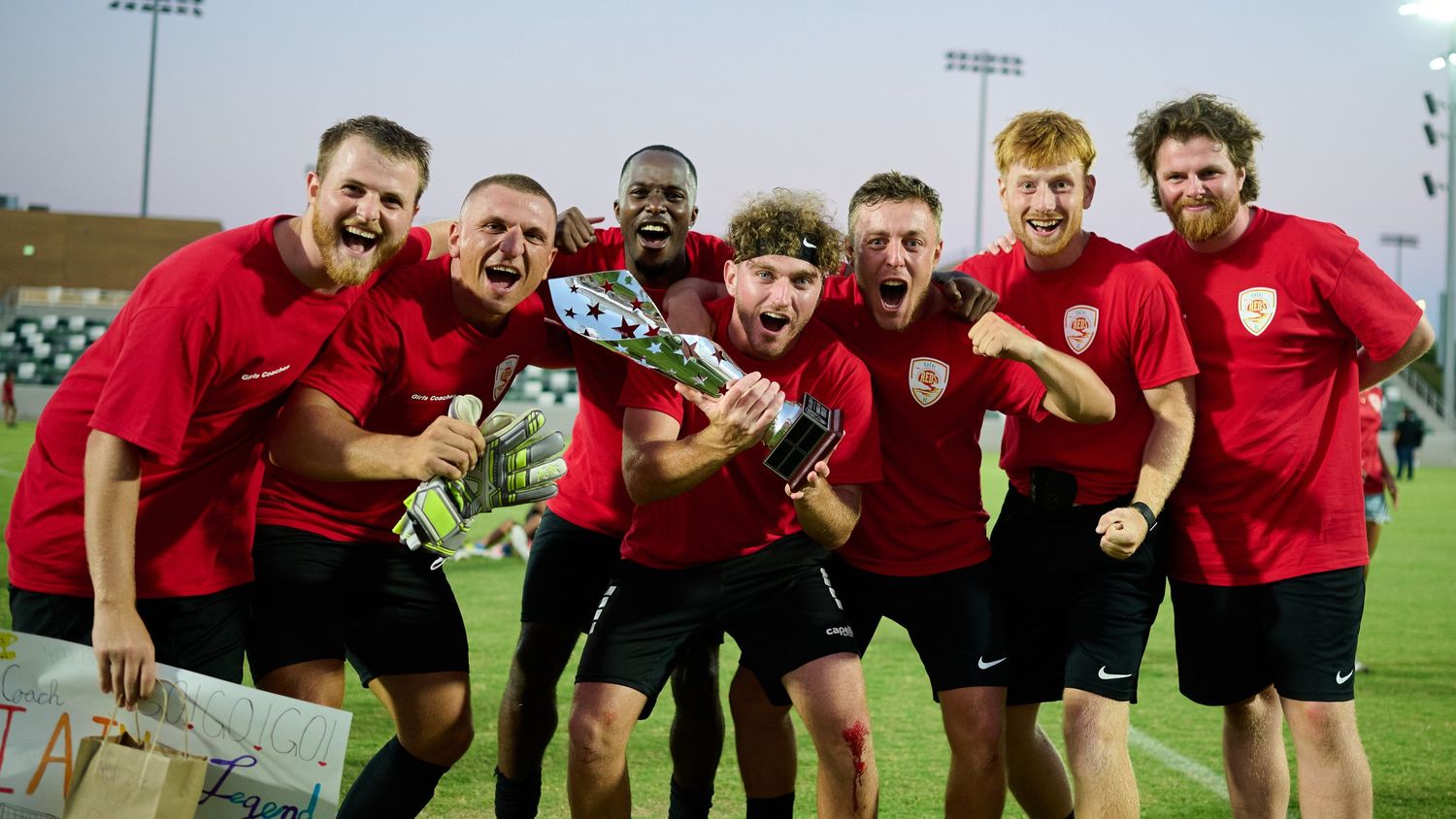 A group of soccer players in red jerseys celebrate together on a field at night.