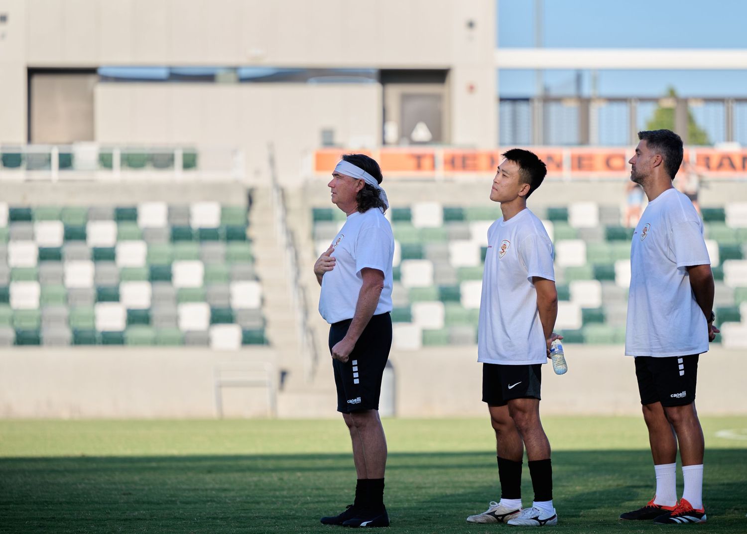 Three soccer players in white jerseys stand in a row on a field during training at an outdoor stadium.