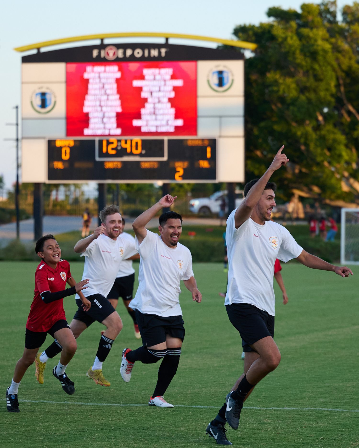Soccer players leap in synchronized motion during a defensive wall formation at a field with scoreboard visible.