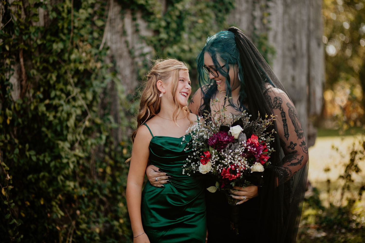 New bride in black wedding dress and veil embraces younger sister in a dark green bridesmaid's dress, barn wall in background, Richmond KY