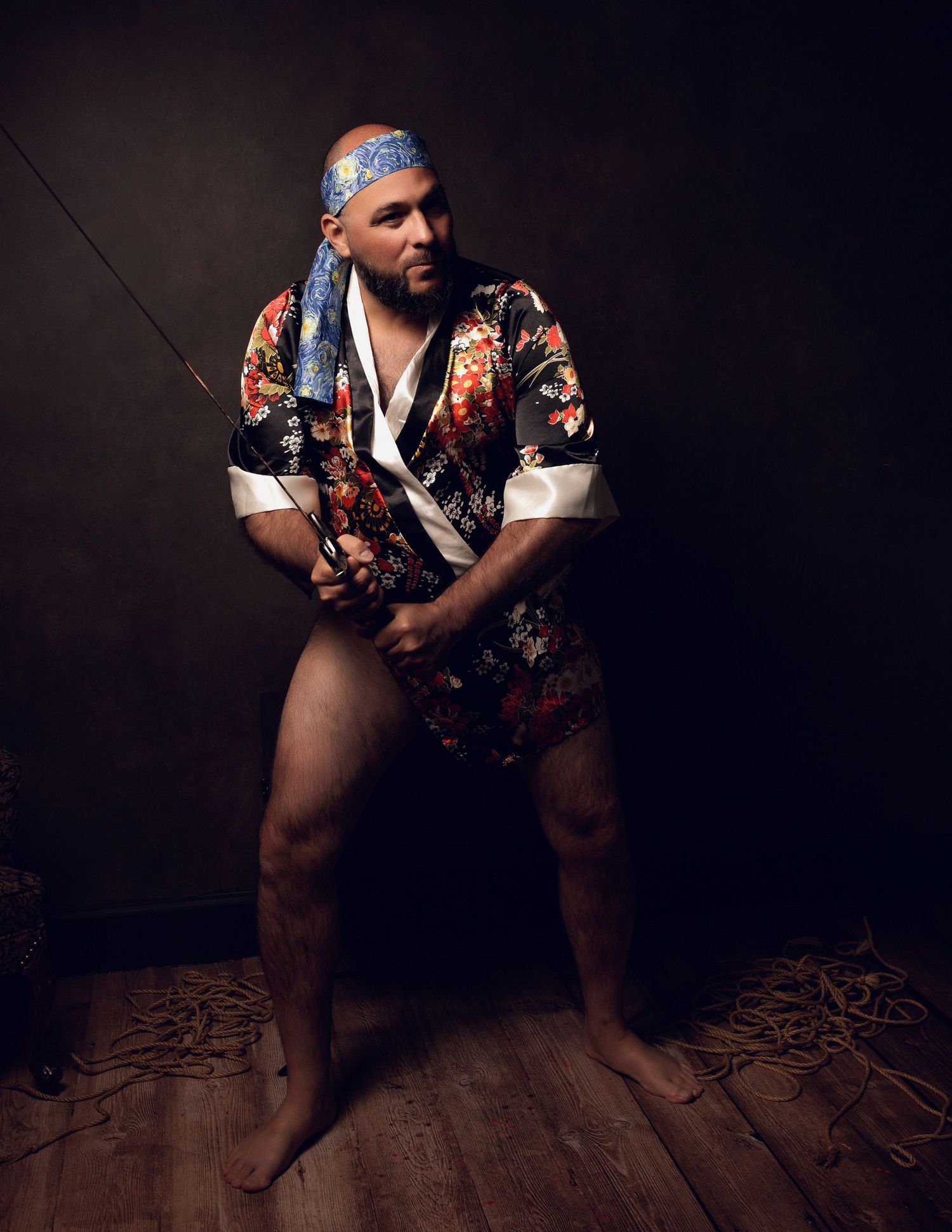Dramatic low-angle portraits on wooden floor wearing floral shirt and bandana in dark setting.