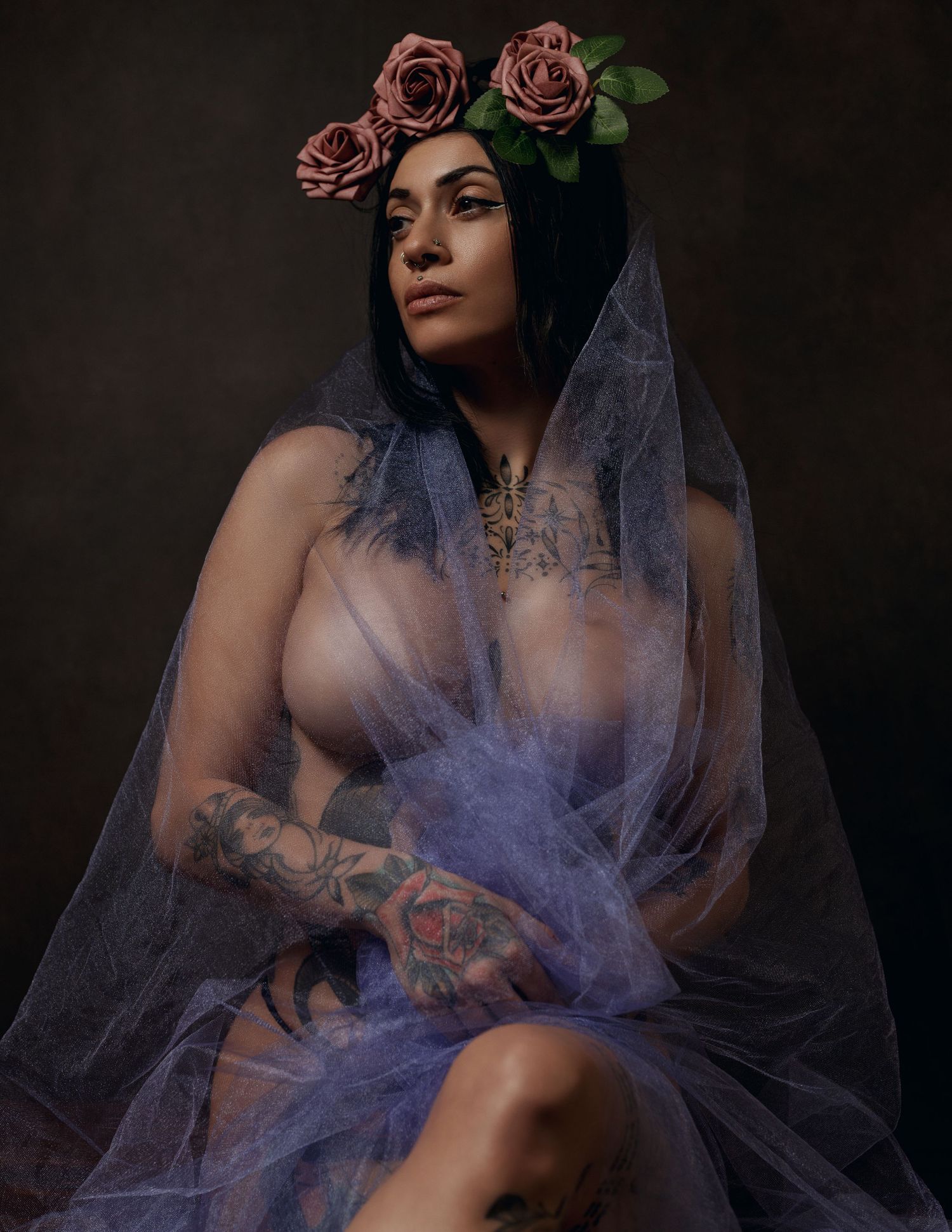 A moody boudoir photoshoot with dark lighting features flowing purple tulle fabric and pink roses as decorative elements.