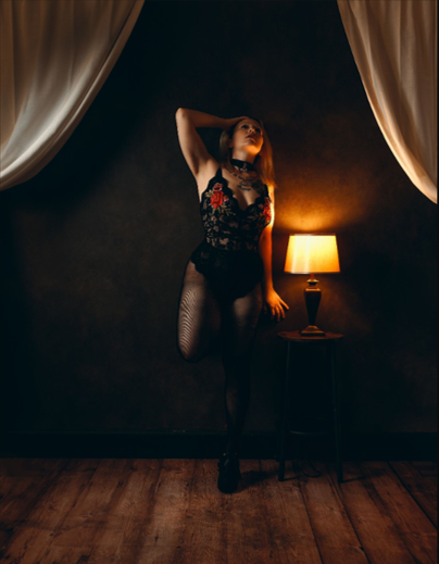 Dramatic boudoir photo in dark room with warm lamp light illuminating figure in black lace against curtained backdrop.