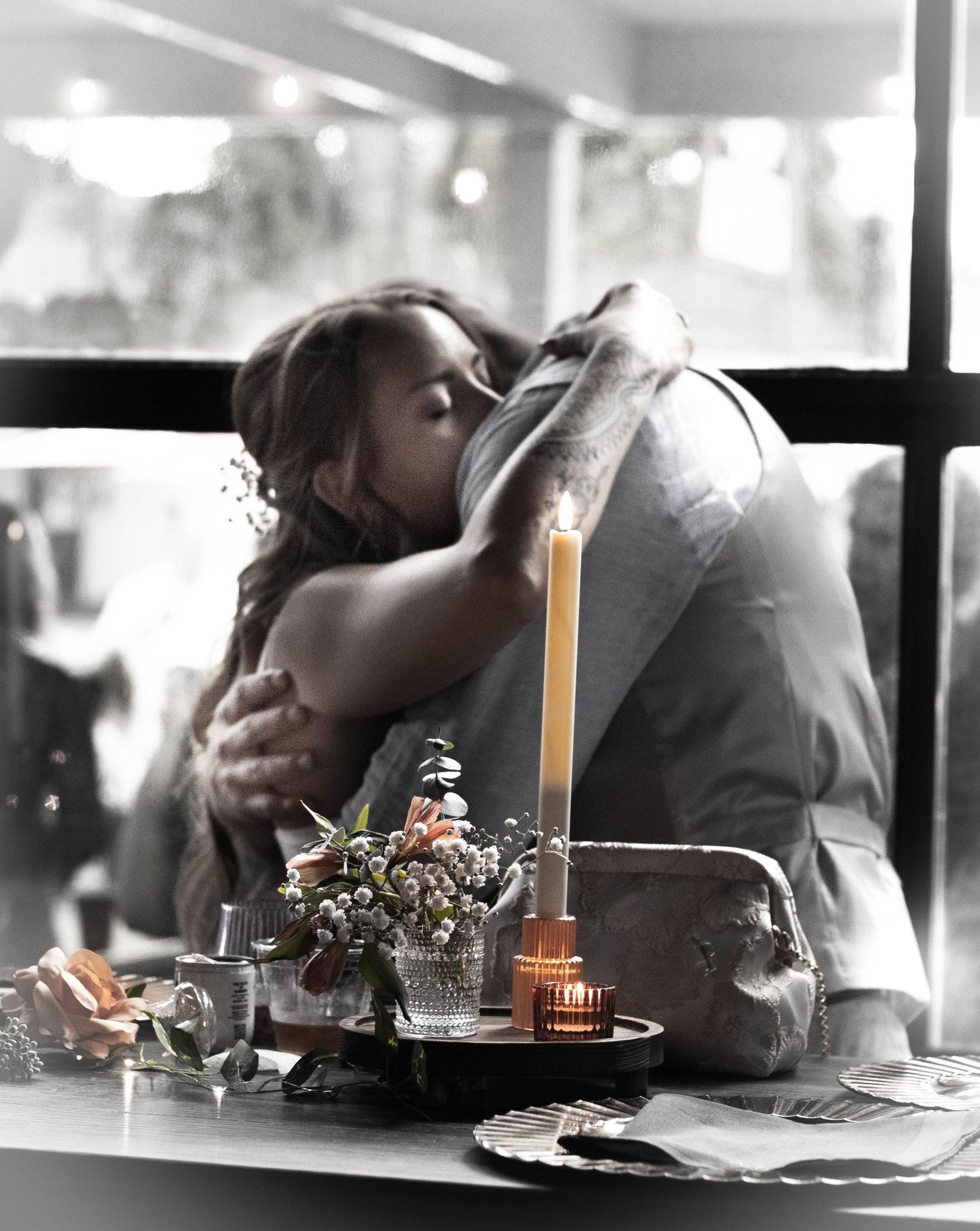 Romantic couple embracing at candlelit dinner table with flowers, candle, and soft bokeh background.