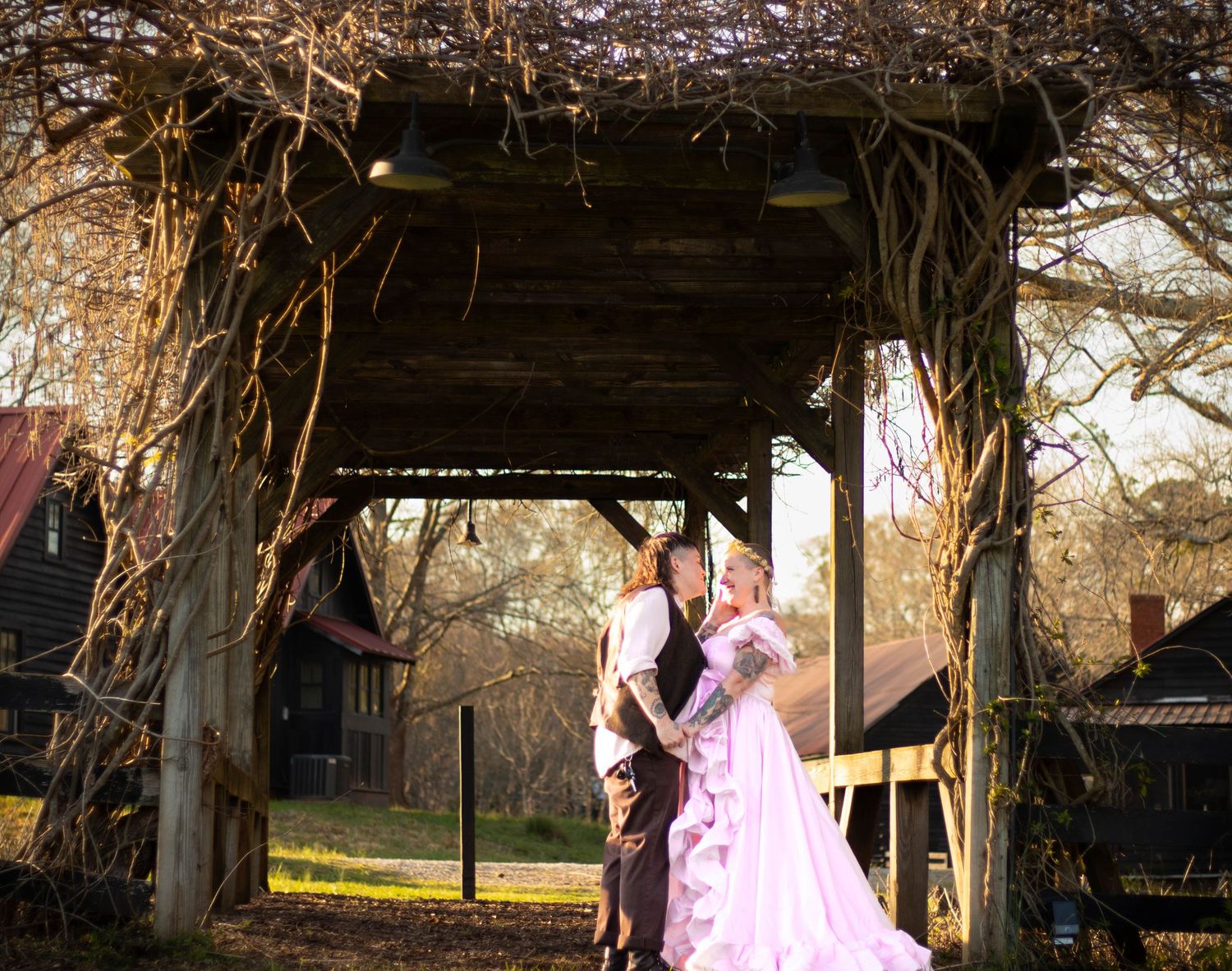 Couple in Renaissance costumes pose under rustic vine-covered pergola in golden hour light.