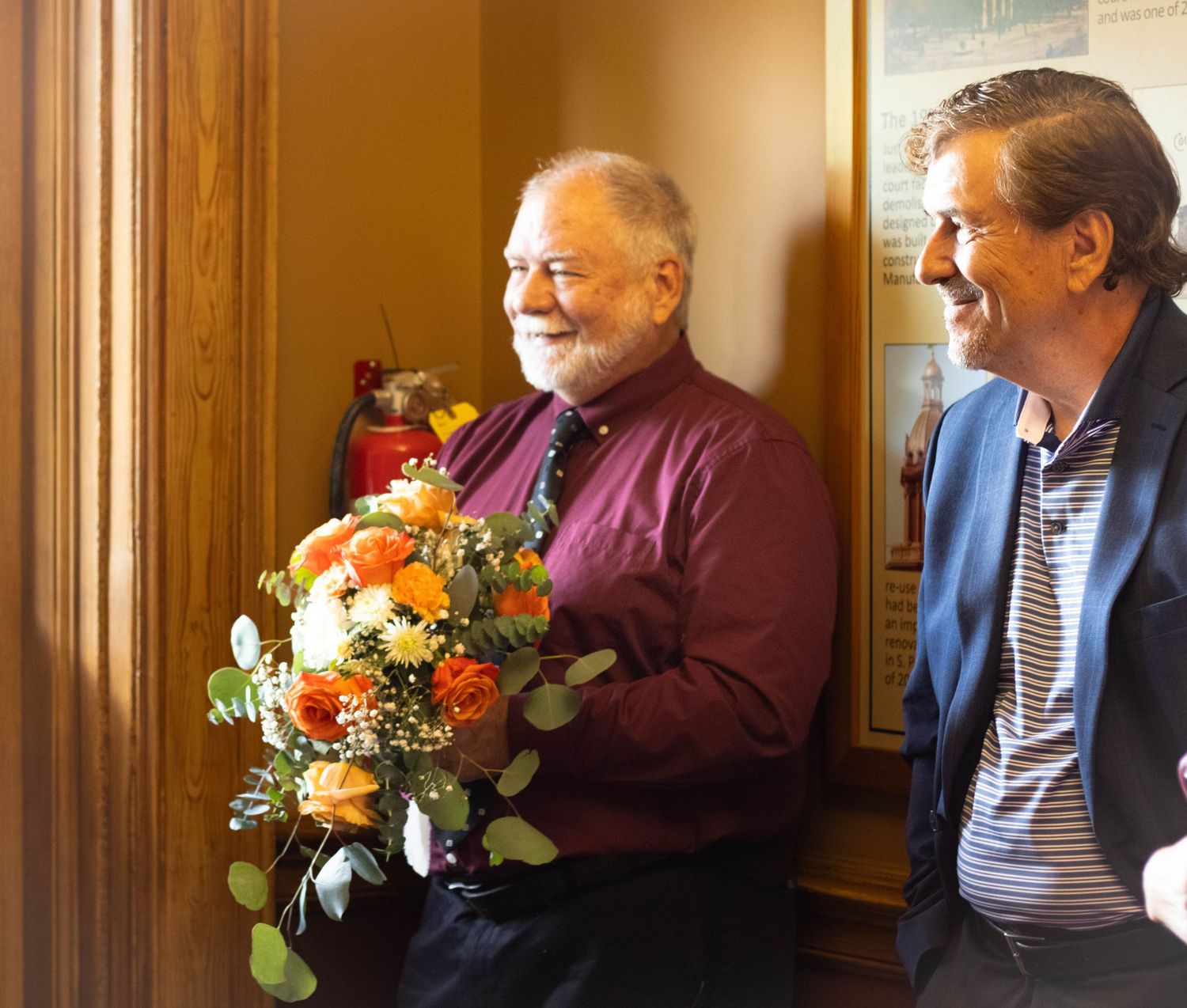 Two smiling men stand indoors, one holding a vibrant bouquet of orange and yellow flowers.
