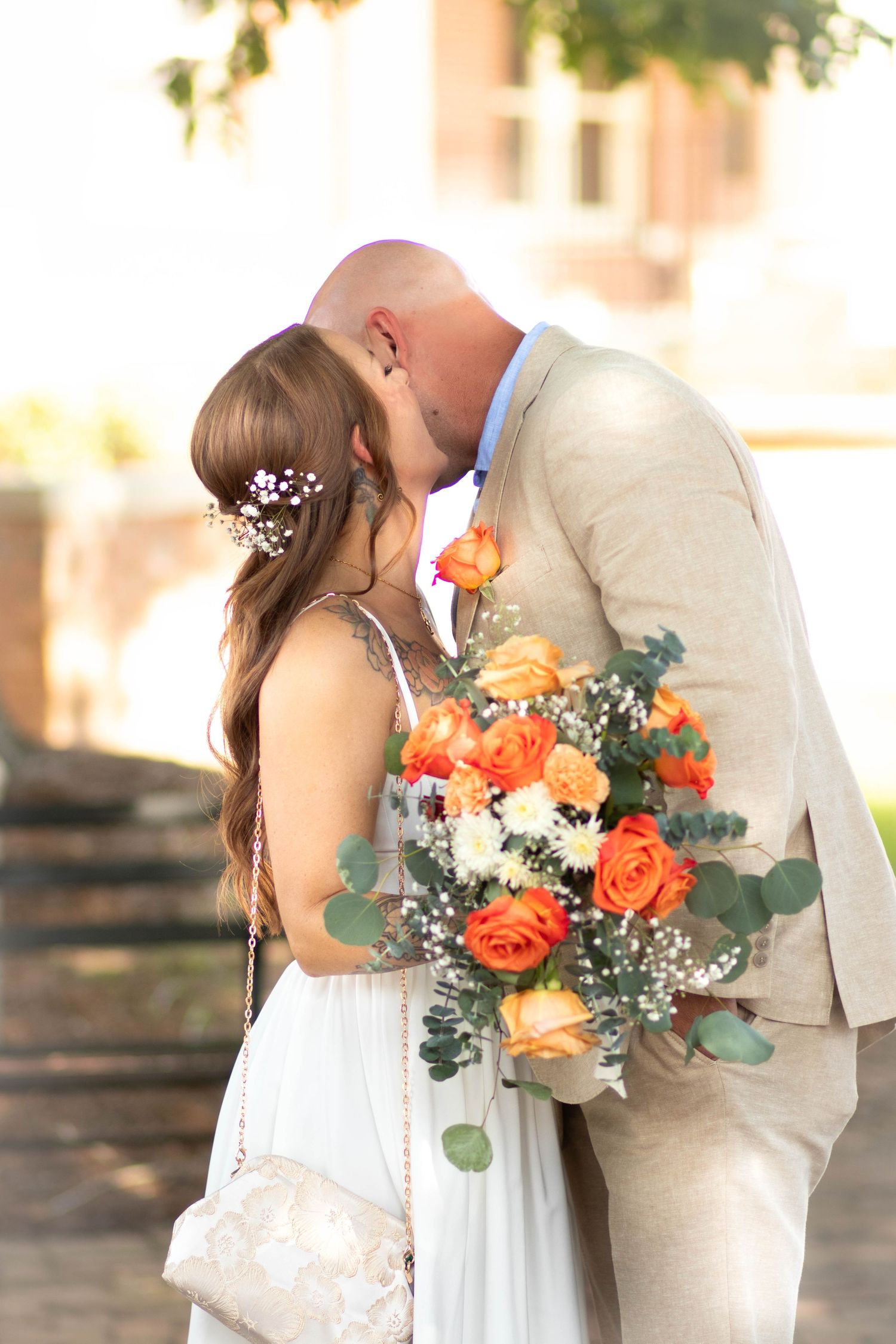 Bride and groom share a kiss outdoors, bride holding vibrant orange and white rose wedding bouquet.