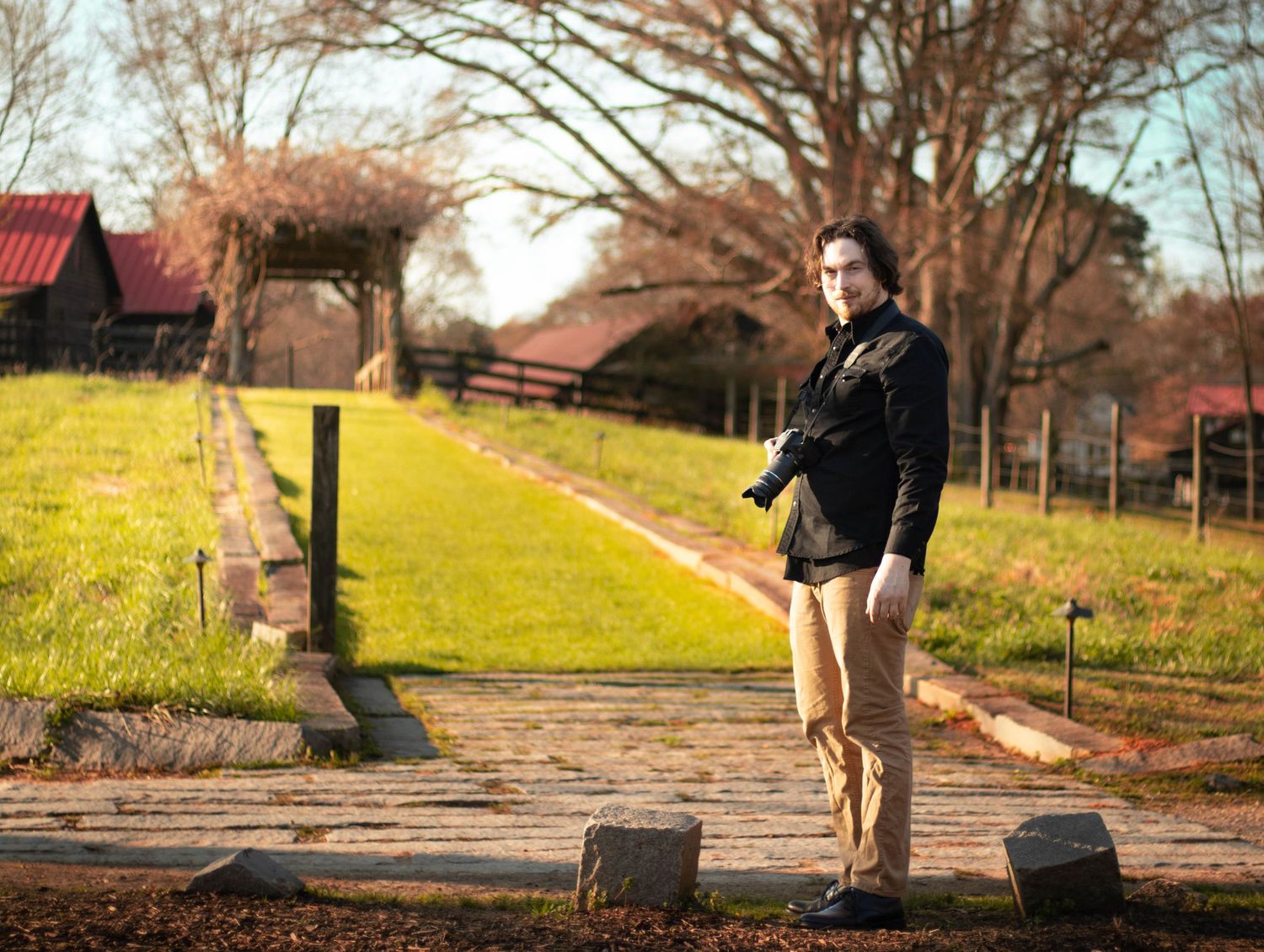 Photographer standing on stone path at rustic farm holding camera during golden hour.