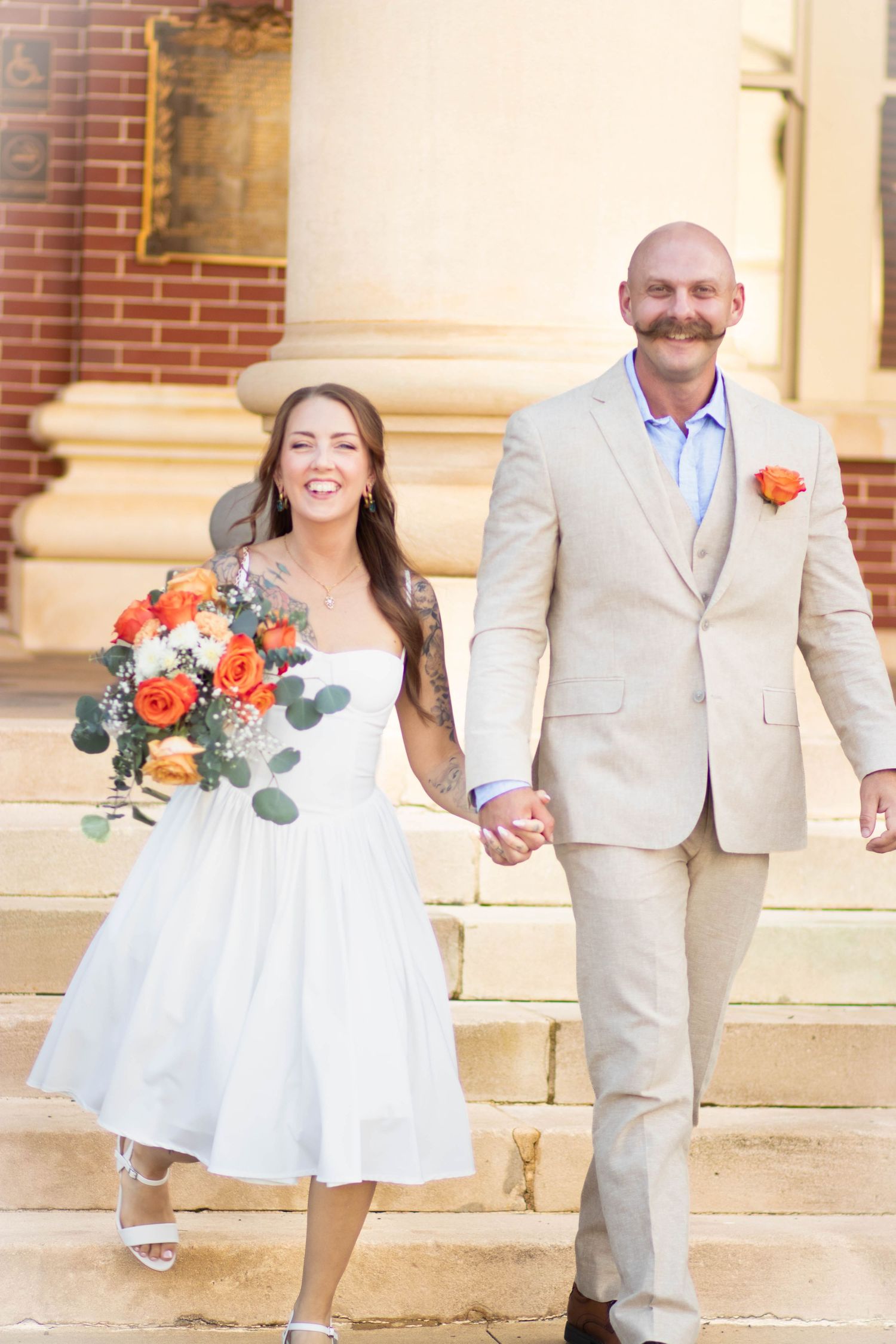 Smiling bride in white dress holding orange bouquet walks hand-in-hand with groom in tan suit on stone steps.